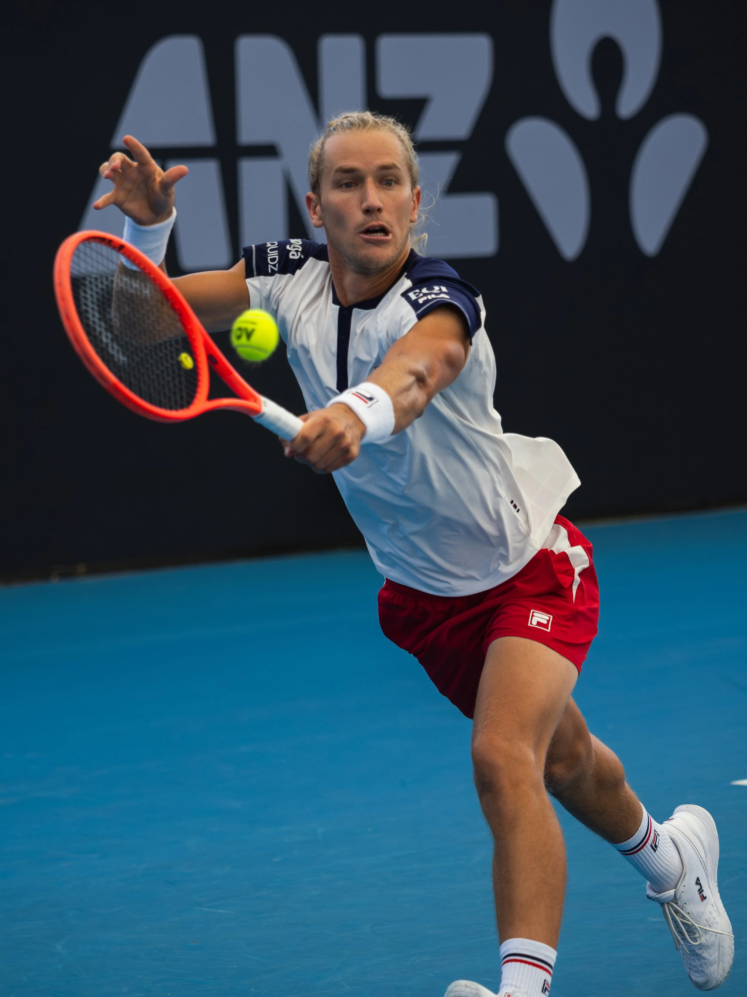A male tennis player with long blonde hair tied back, wearing a white and navy sports shirt, red shorts, and white tennis shoes, is hitting a tennis ball with a bright orange racket on a blue tennis court.