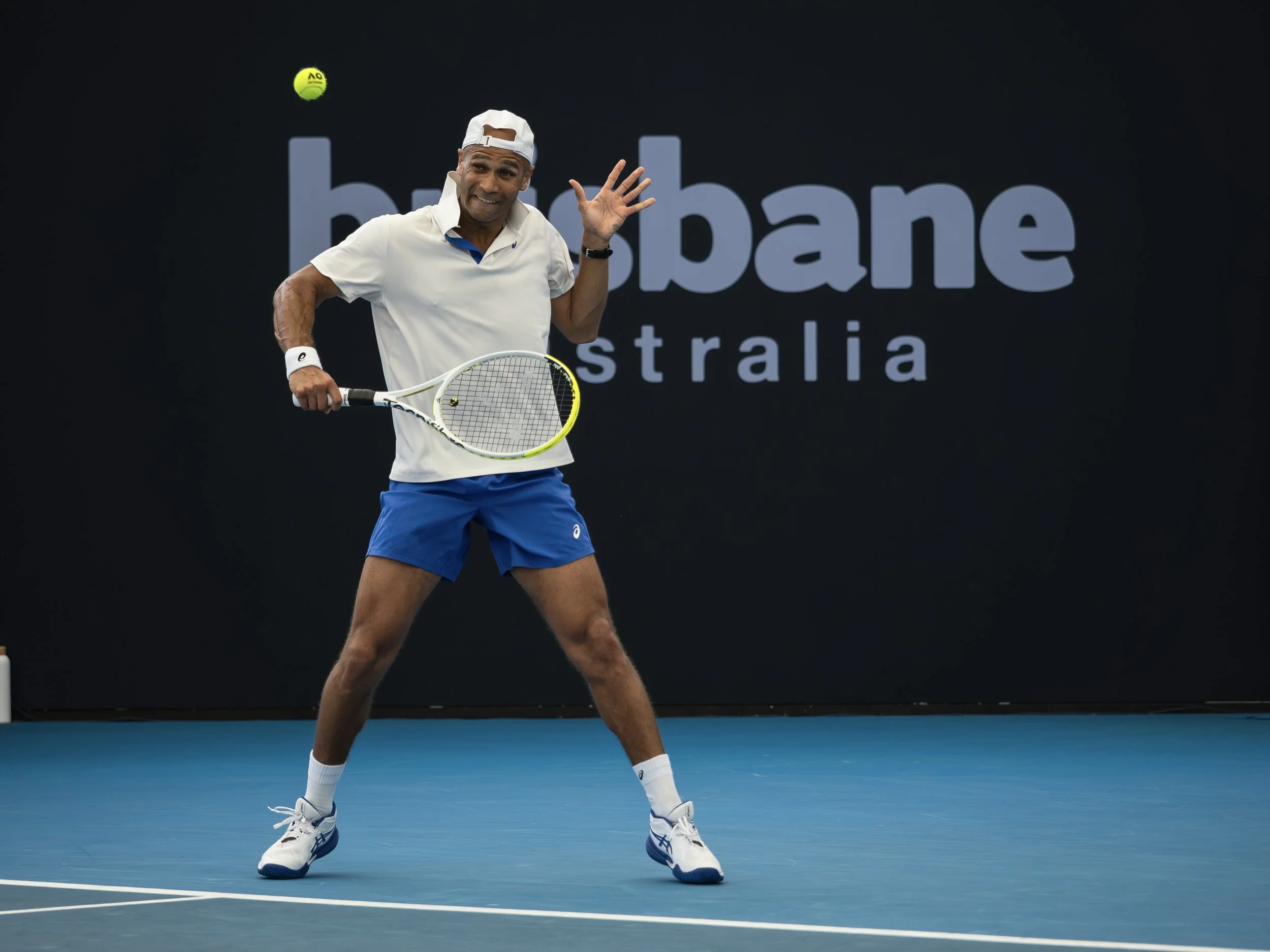 Tennis player celebrating on the court with a smile, holding a tennis racket, wearing a white cap, white shirt, blue shorts, and white shoes, with a black background and Australian Open signage.