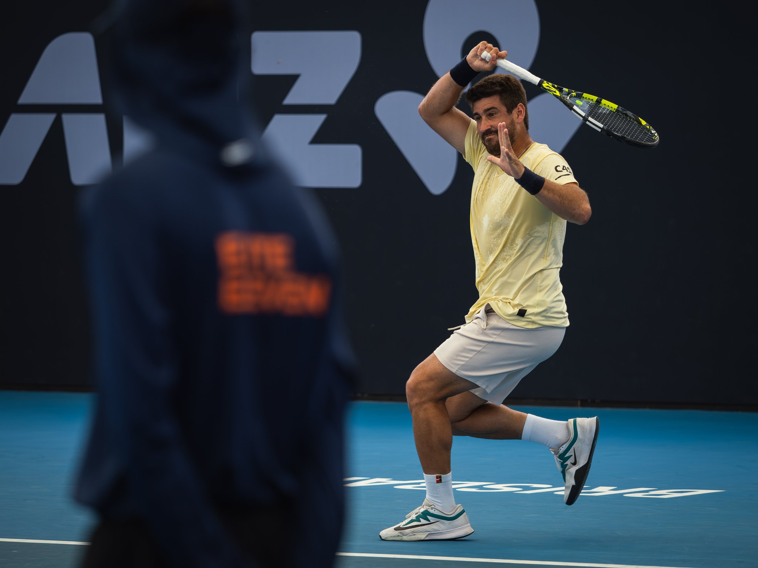 A male tennis player in a yellow shirt and white shorts is on a blue tennis court, with a black background and large white sponsor logos. He is preparing to serve, holding a tennis racket and ball, with one knee on the ground and the other leg bent.