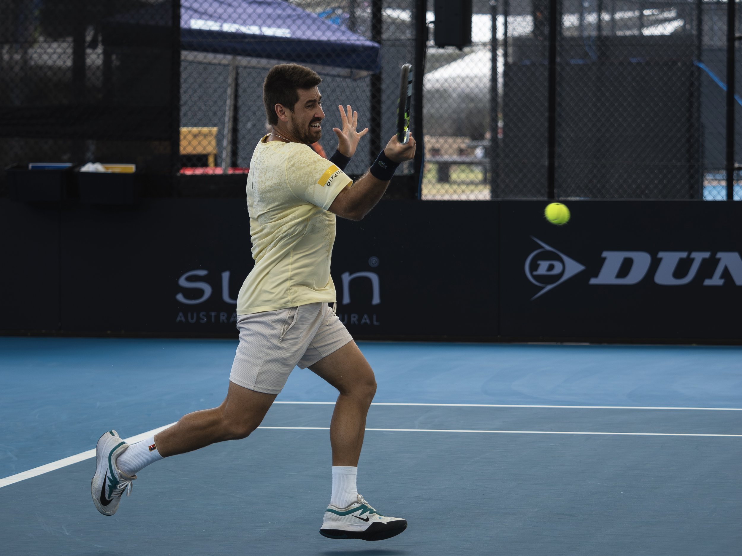 A male tennis player running on a blue tennis court with a green tennis ball in mid-air in front of him. He is wearing a yellow shirt, white shorts, white socks, and sneakers, with a wristband on his right arm and a necklace. The background shows a b