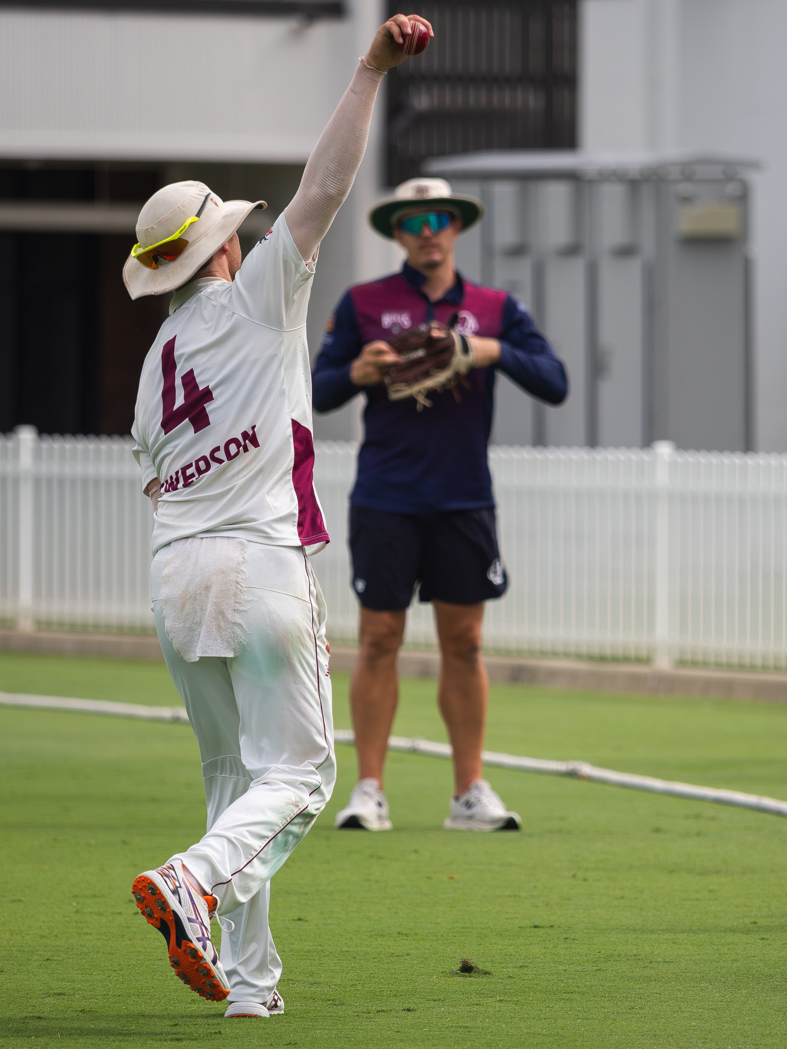 A cricket player in white uniform and wide-brimmed hat is bowling a ball on a cricket field; another person in sports attire and sunglasses is in the background.