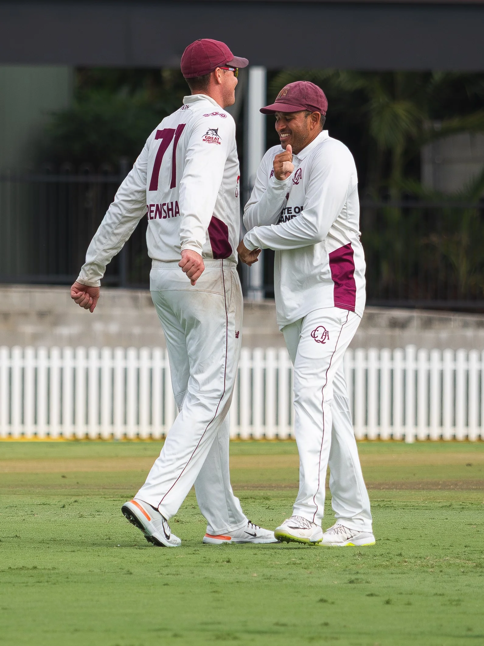 Two cricket players, wearing white uniforms and maroon caps, are walking and laughing on a cricket field, with a white picket fence and some greenery in the background.