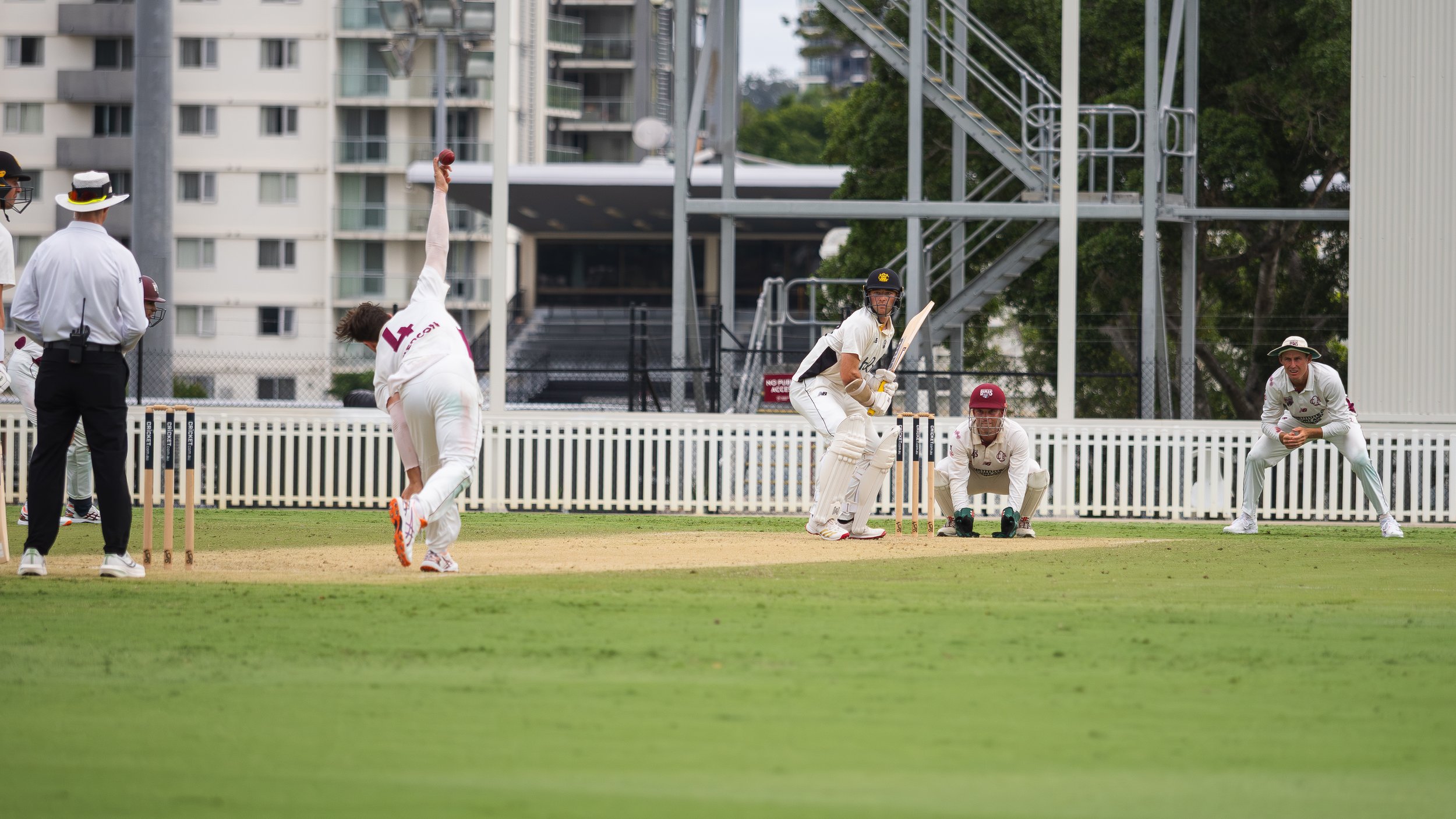 Cricket players on the field during a match, with a batter at the crease and bowlers and fielders in position, with a background of buildings and trees.