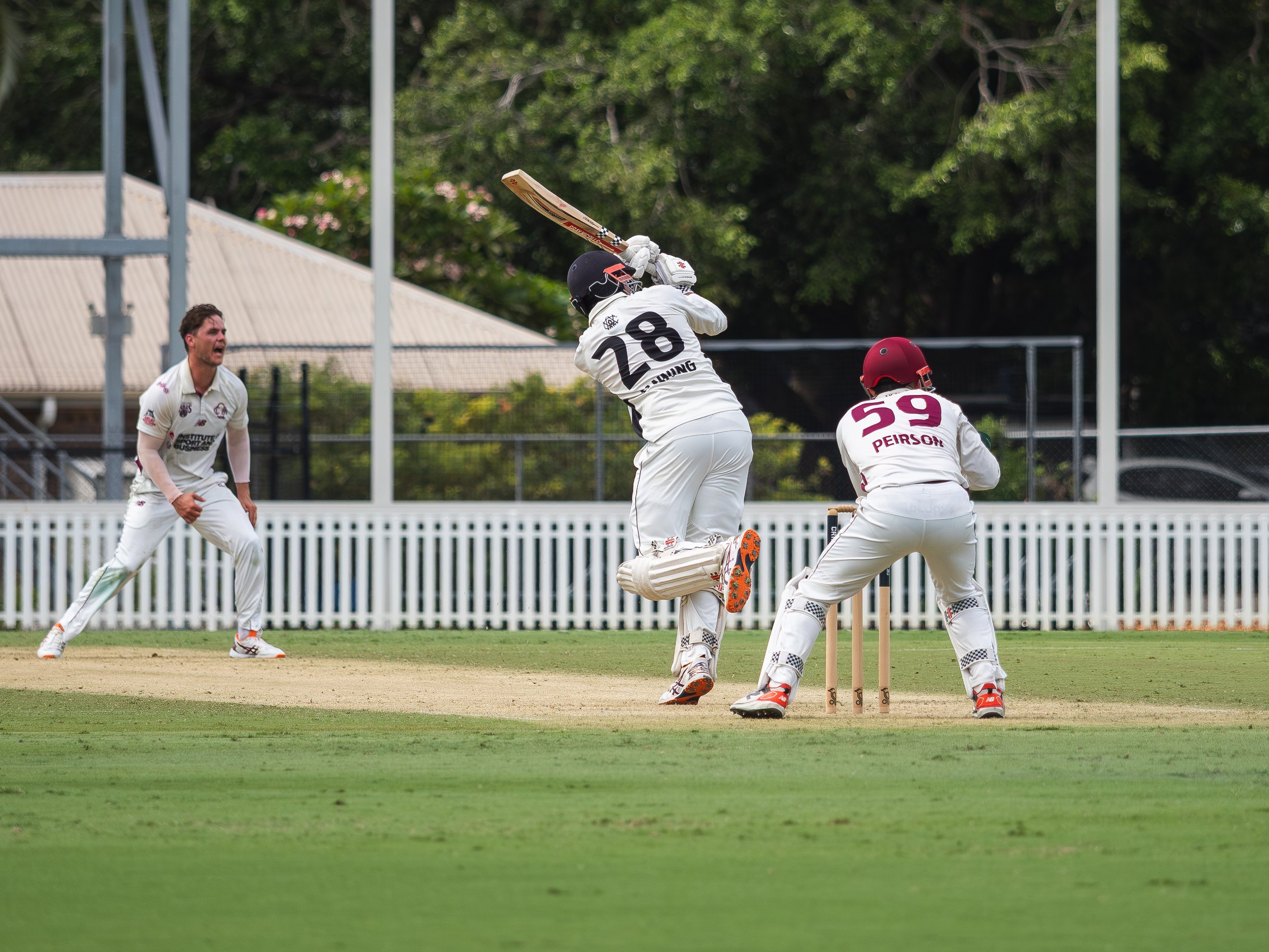 Cricket players on a field, with one player preparing to hit the ball, while others are positioned around. The players are wearing white uniforms, and there are trees and a fence in the background.