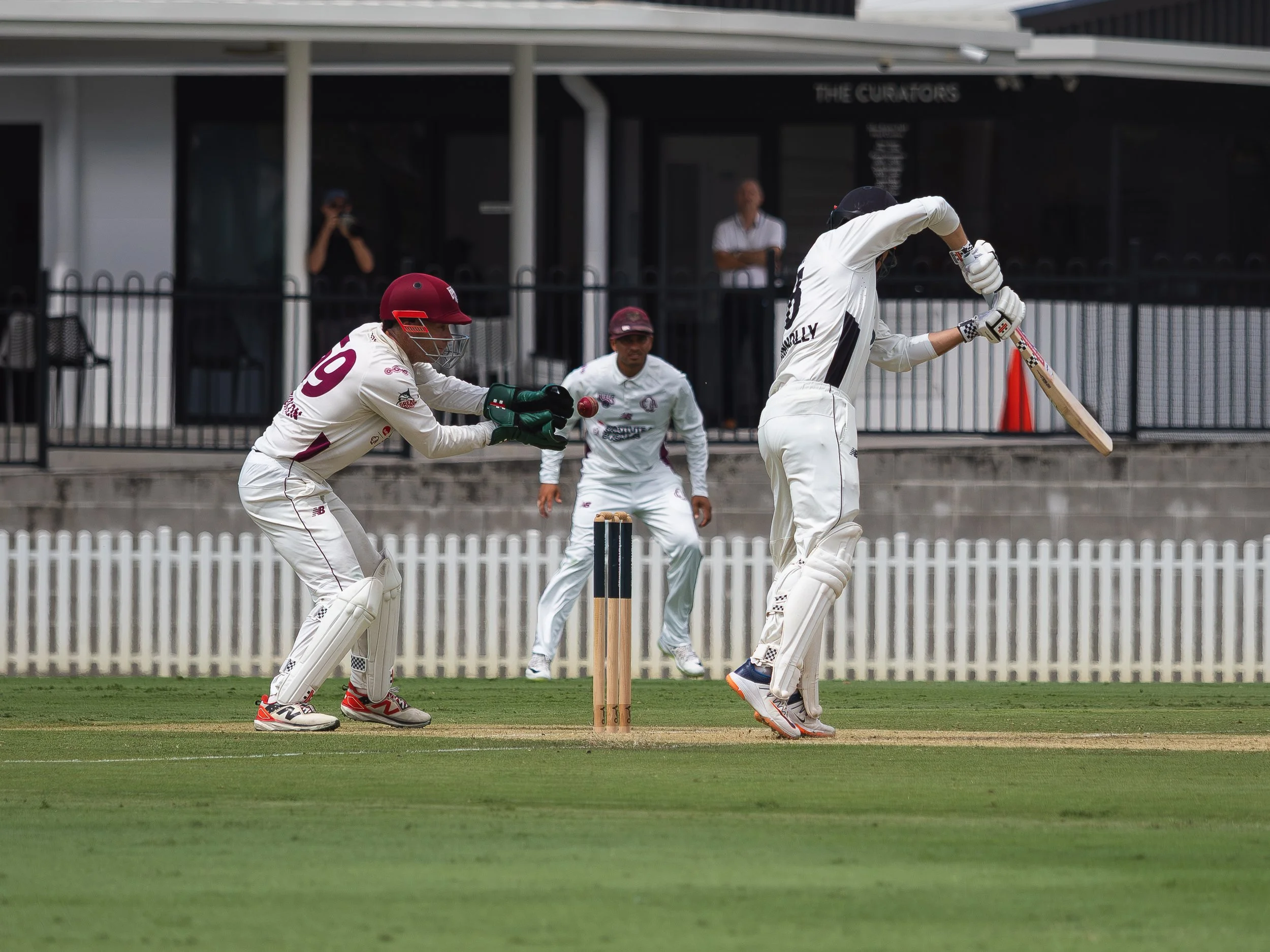 Cricket players on the field, one batting and one wicket-keeping, with a fielder and an umpire nearby, under a stadium roof. The ground is green and the players are in white uniforms.