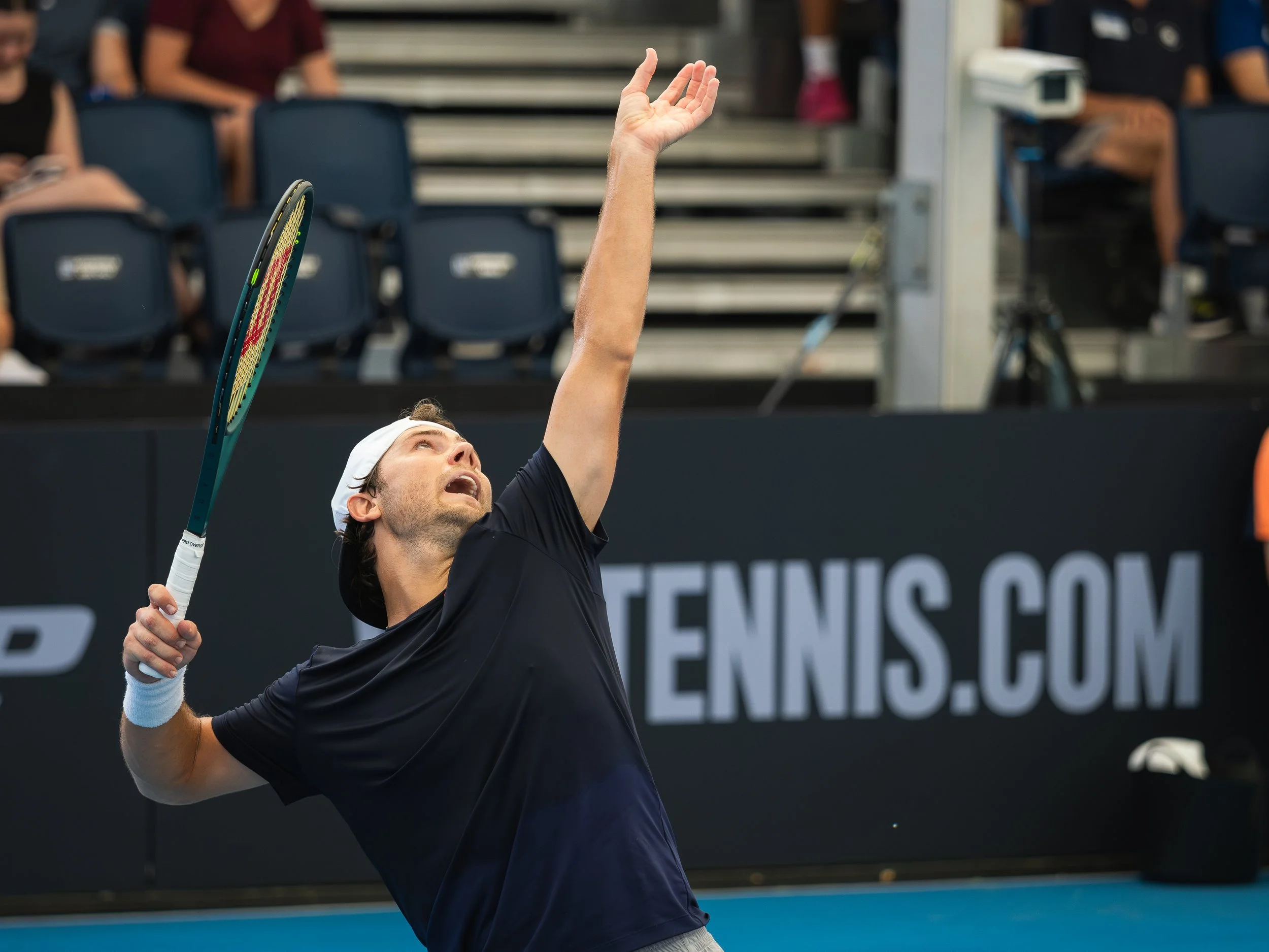 A male tennis player, wearing a black shirt and white cap, is reaching up with his left hand while holding a tennis racquet in his right hand, in a tennis stadium during a match.