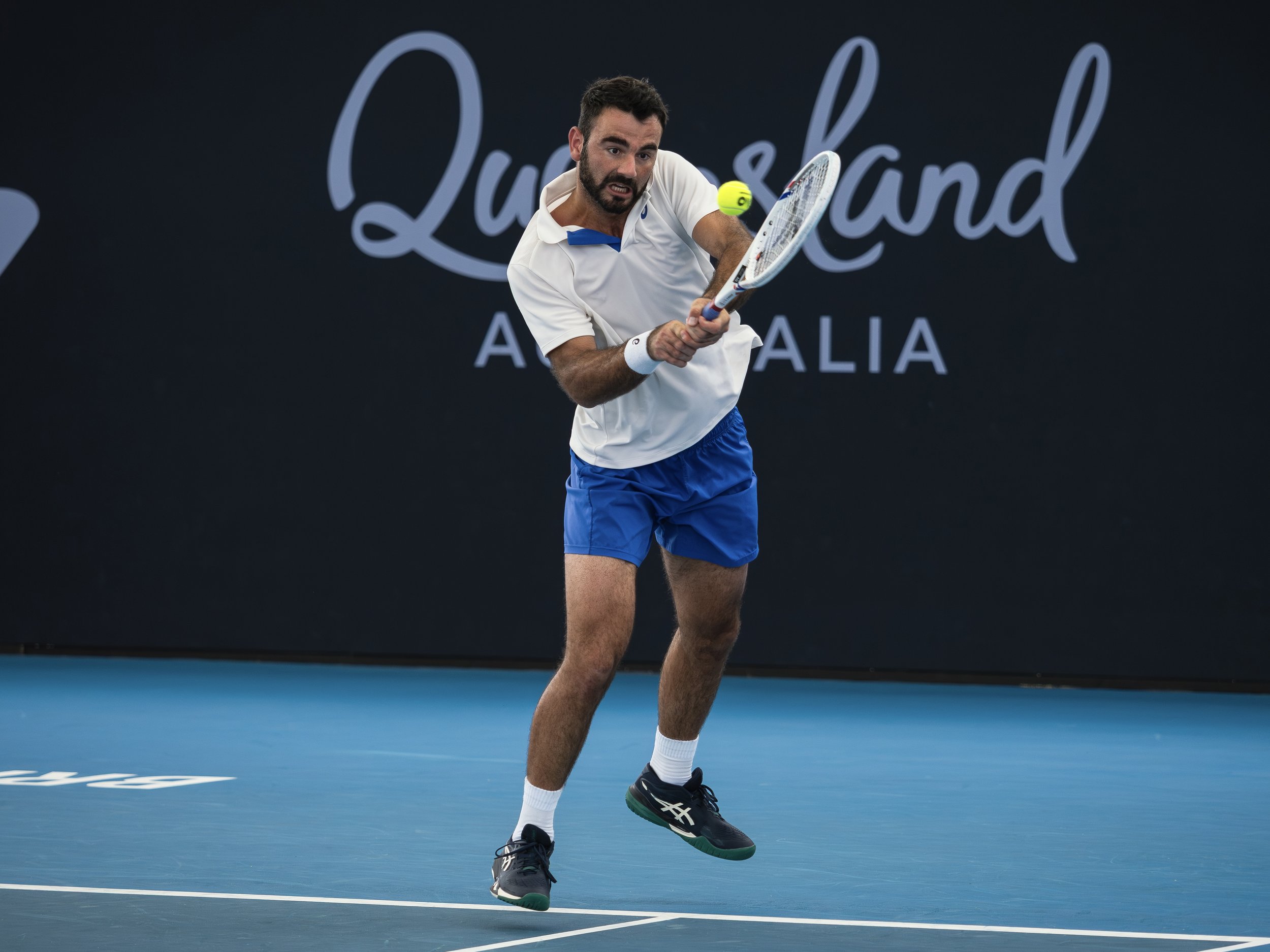 Male tennis player in white shirt and blue shorts hitting a tennis ball with a racket on a blue tennis court.
