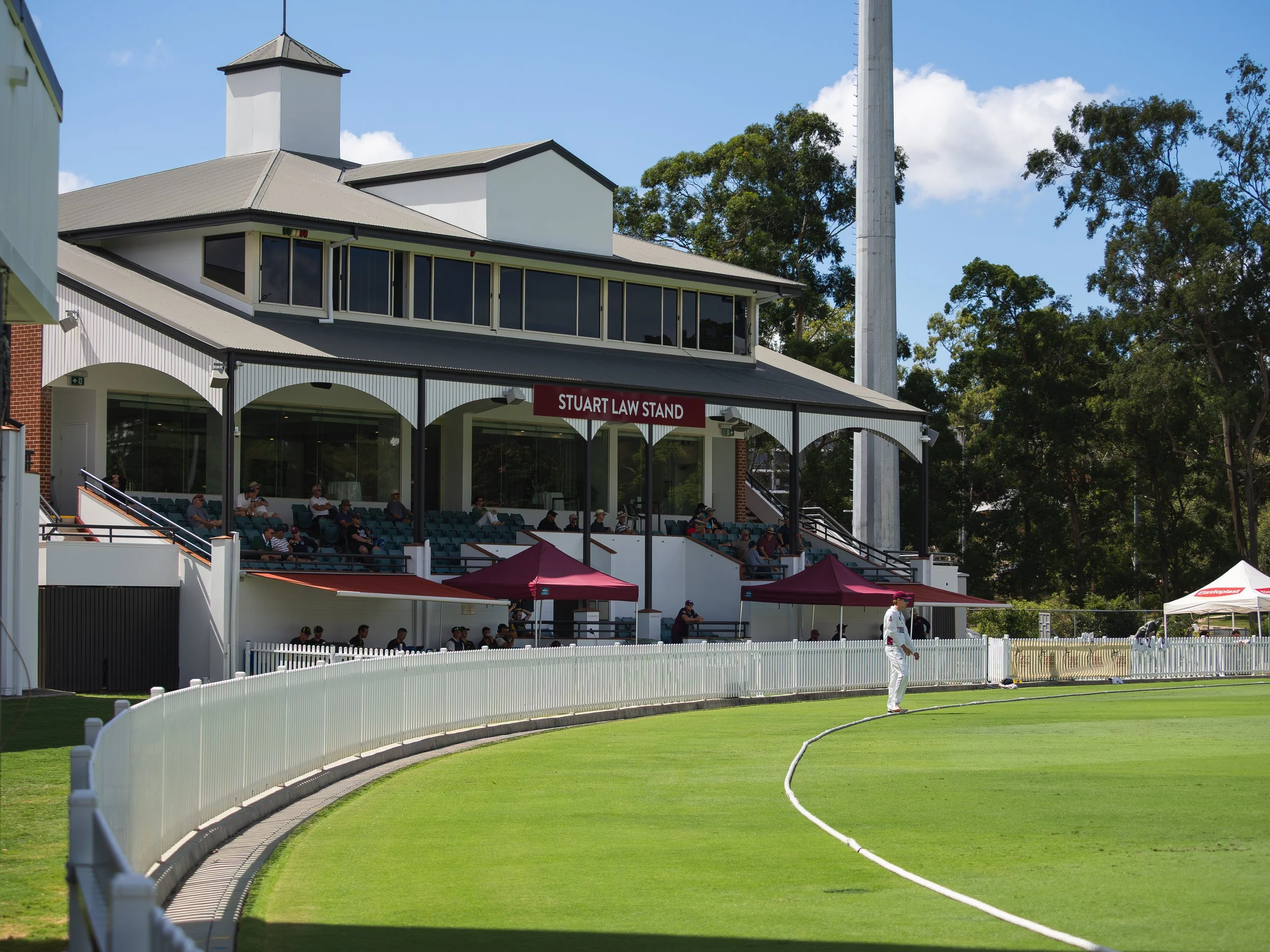 View of a cricket pitch with a player practicing near the pitch, a grandstand with spectators, and a building labeled 'Stuart Law Stand' in the background, surrounded by trees and a blue sky.