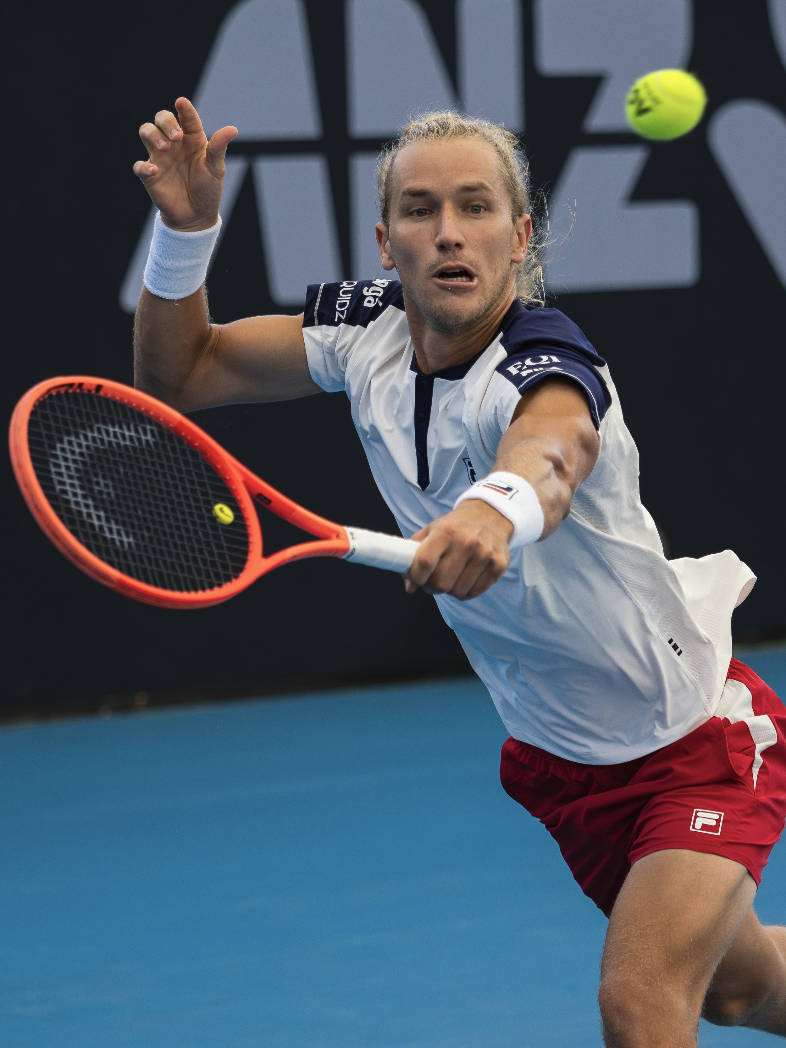 A male tennis player with long blonde hair is in the middle of a forehand shot on a blue tennis court. He is wearing a white and navy sports shirt, red shorts, and a white wristband, with a yellow tennis ball in motion against a black background.