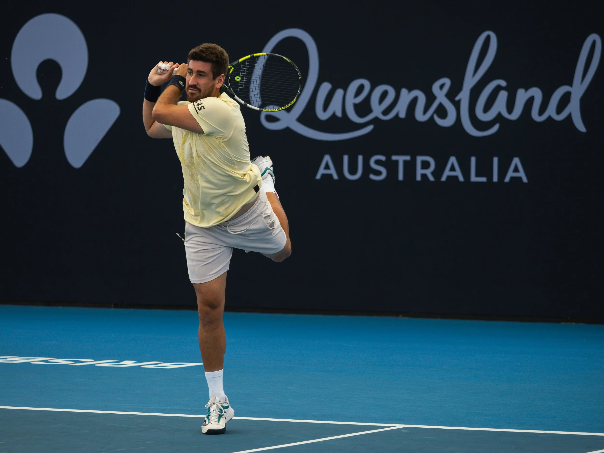 A male tennis player in a yellow shirt and light gray shorts serves on a blue tennis court at the Queensland Australia event.