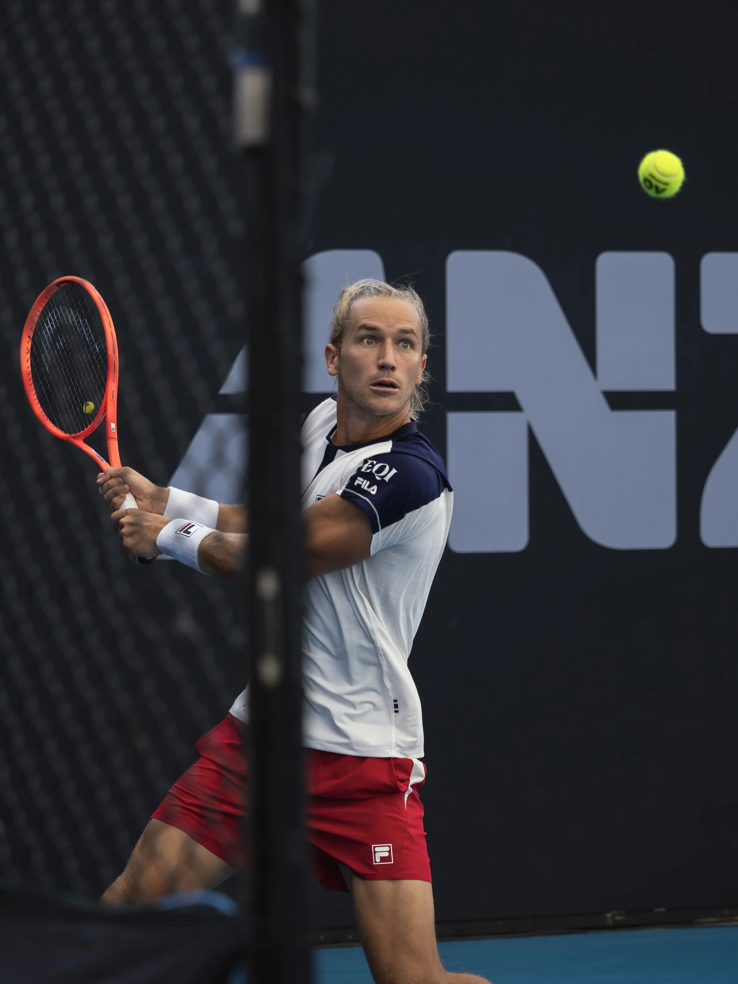 A tennis player with long hair tied back, wearing a white and navy shirt and red shorts, is hitting a tennis ball on a court.