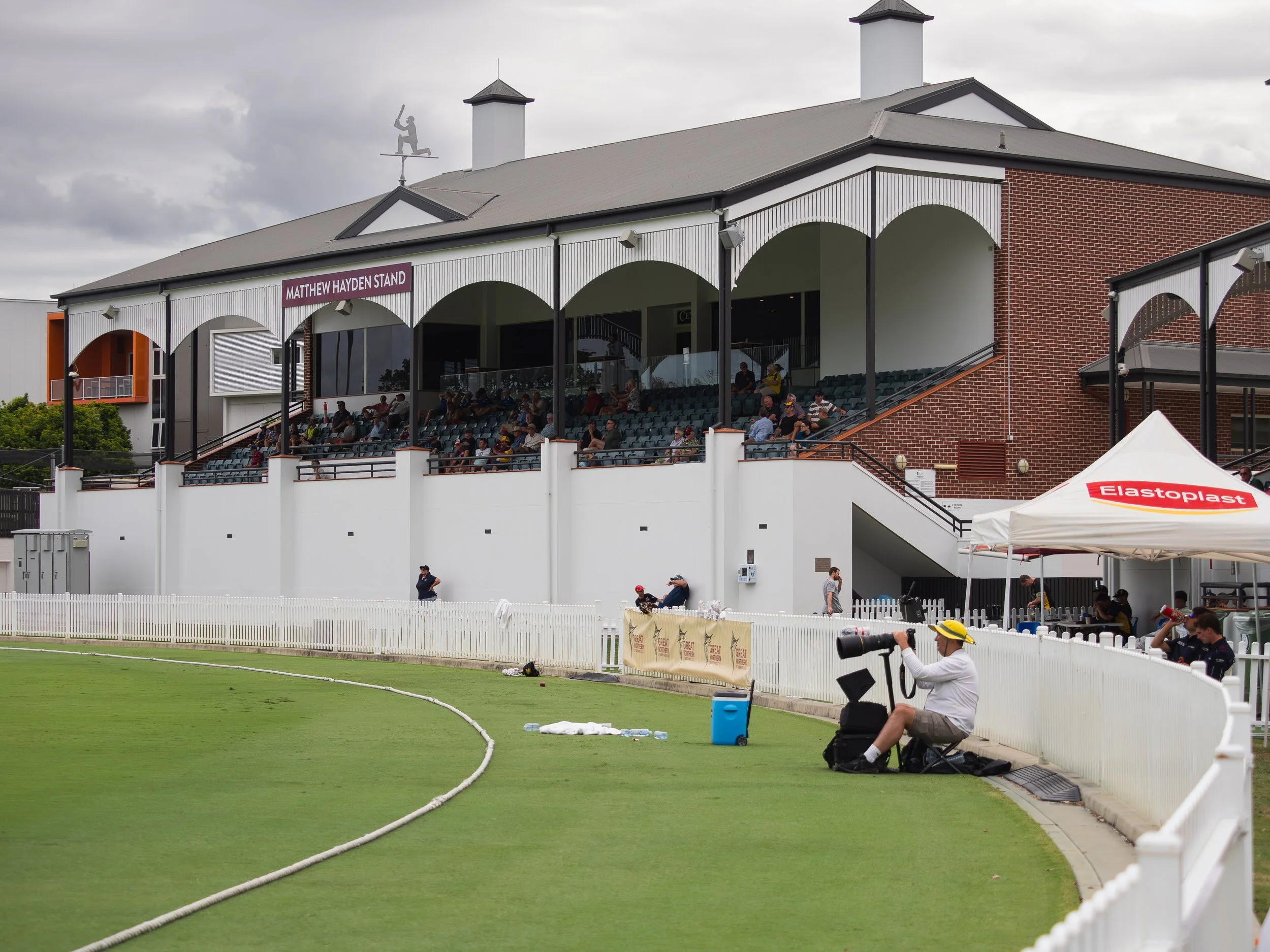 A sports stadium with a seated audience under a covered bleacher area, a manicured grass field, and a person sitting on a foldable chair near the field with a camera and other equipment, during cloudy weather.