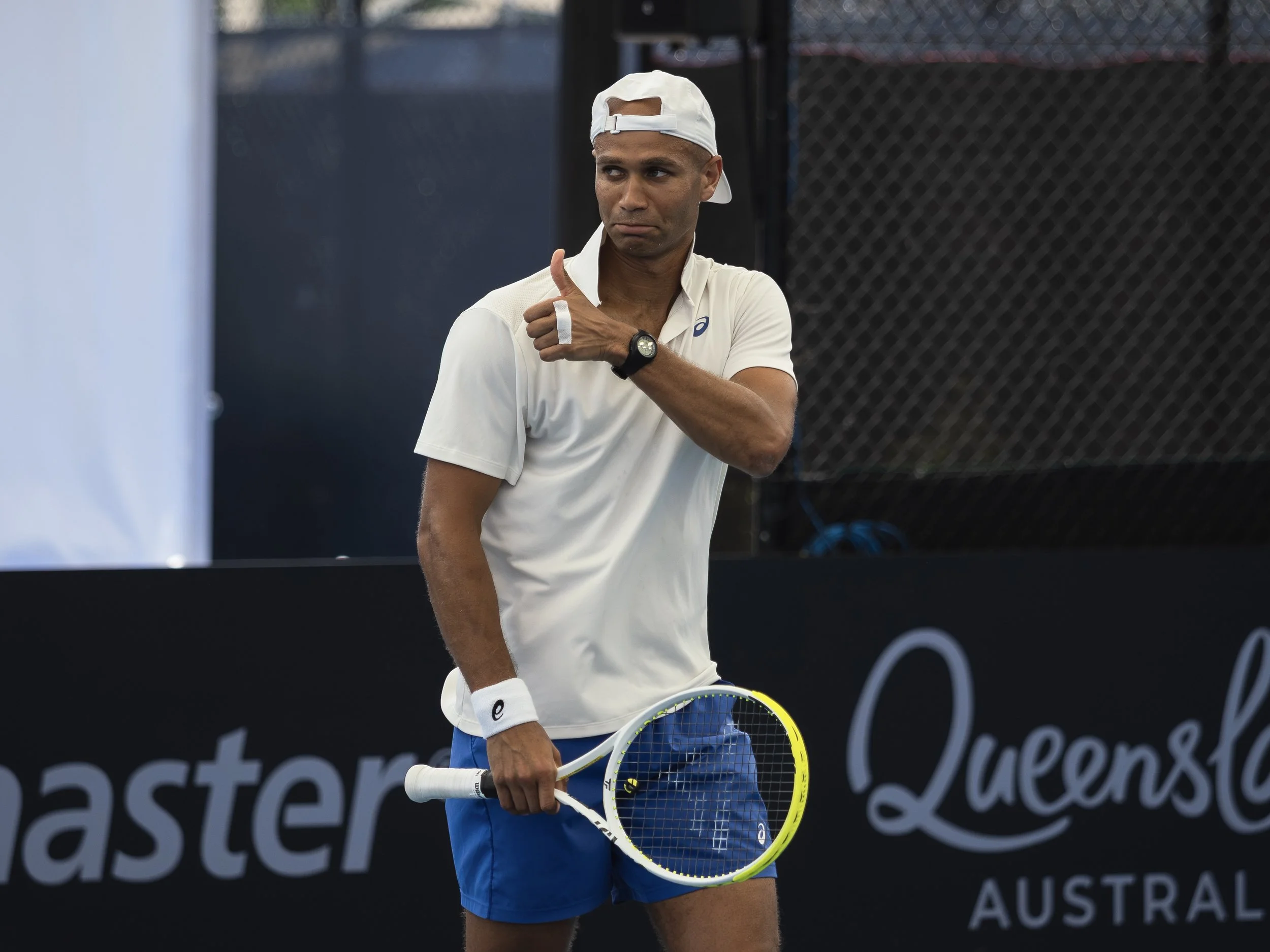 A male tennis player wearing a white cap, white shirt, and blue shorts, holding a tennis racket, standing on a tennis court with a black fence in the background.