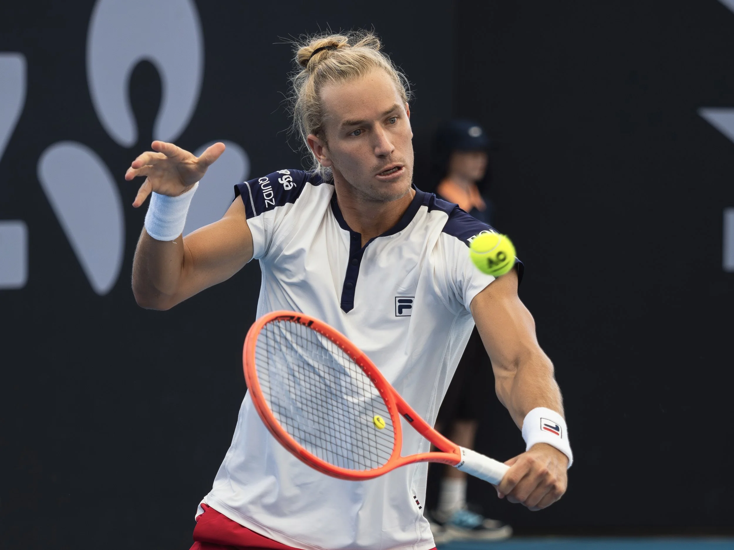 A male tennis player with long blond hair tied in a bun, wearing a white and navy shirt, holding a tennis racket, about to hit a tennis ball.