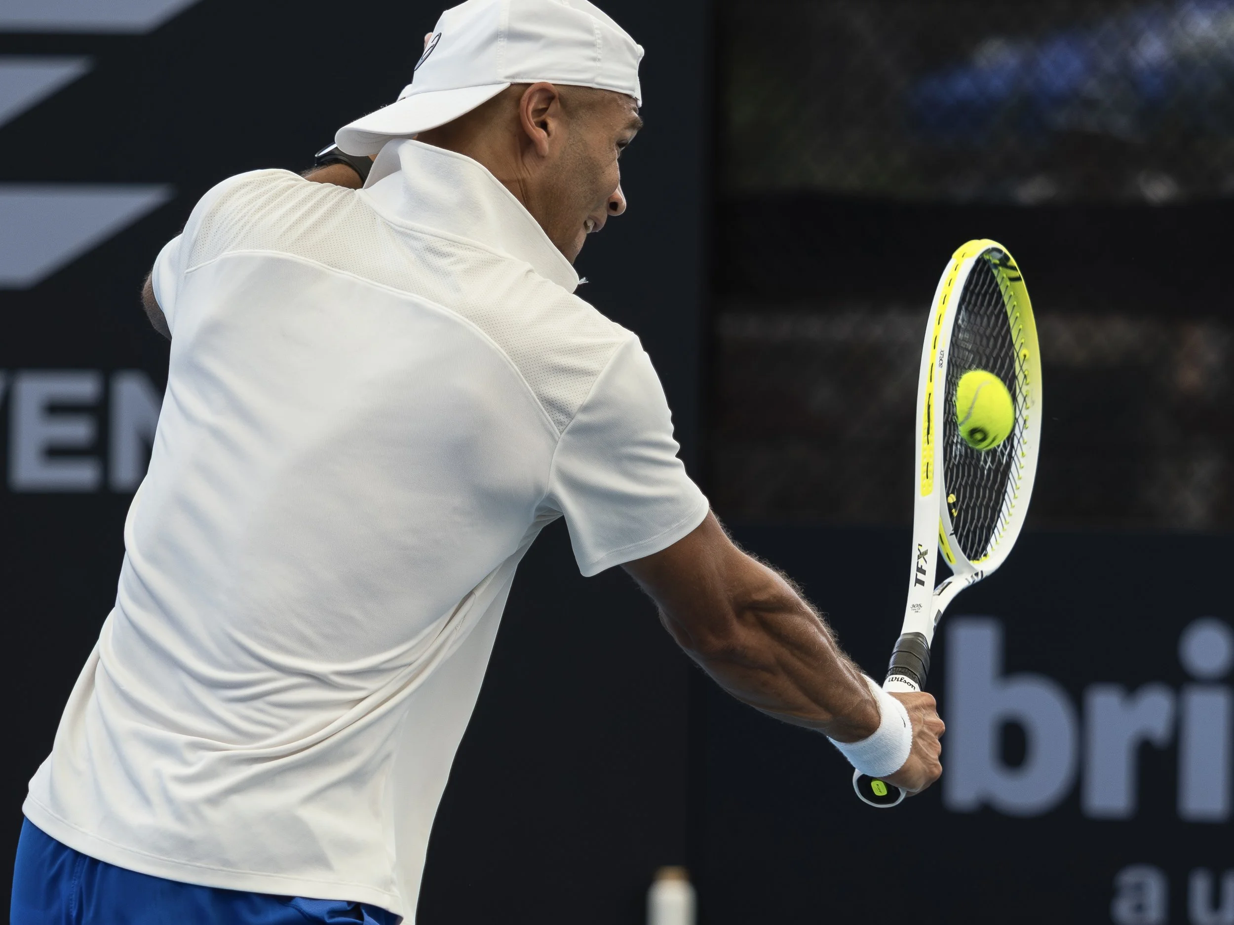 A male tennis player wearing a white cap and shirt, holding a tennis racket with a yellow tennis ball, ready to hit the ball on a tennis court.