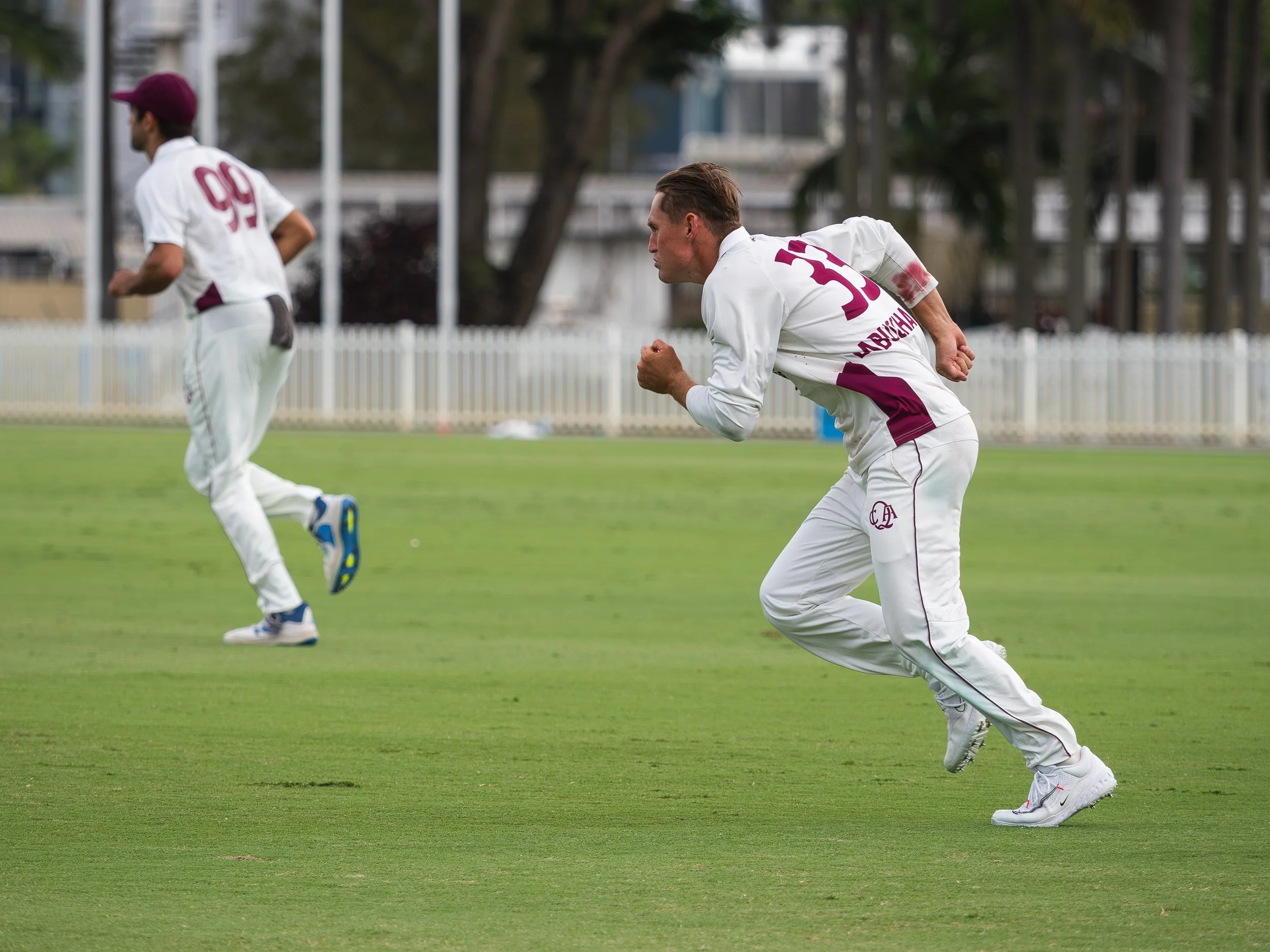 Two cricket players in white uniforms running on a green field during a match, with trees and a fence in the background.