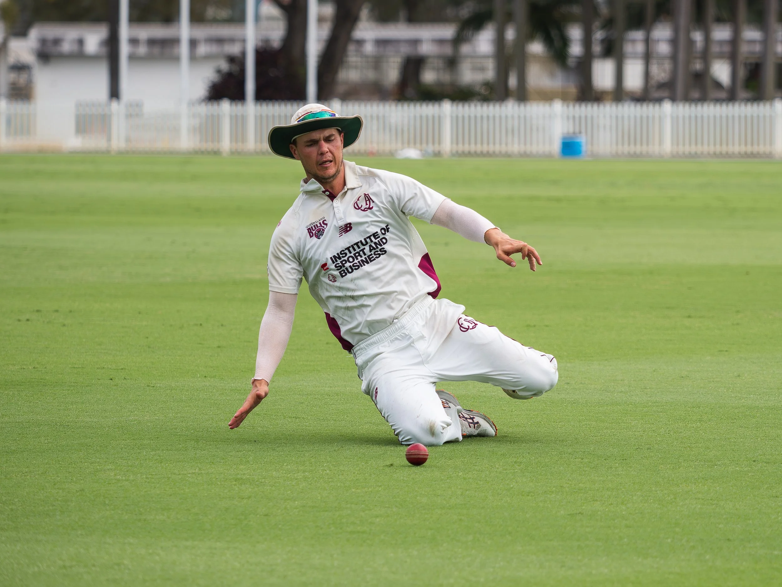 A cricket player falling on the ground during a match, wearing a white uniform with a green hat, on a green field surrounded by a white fence.
