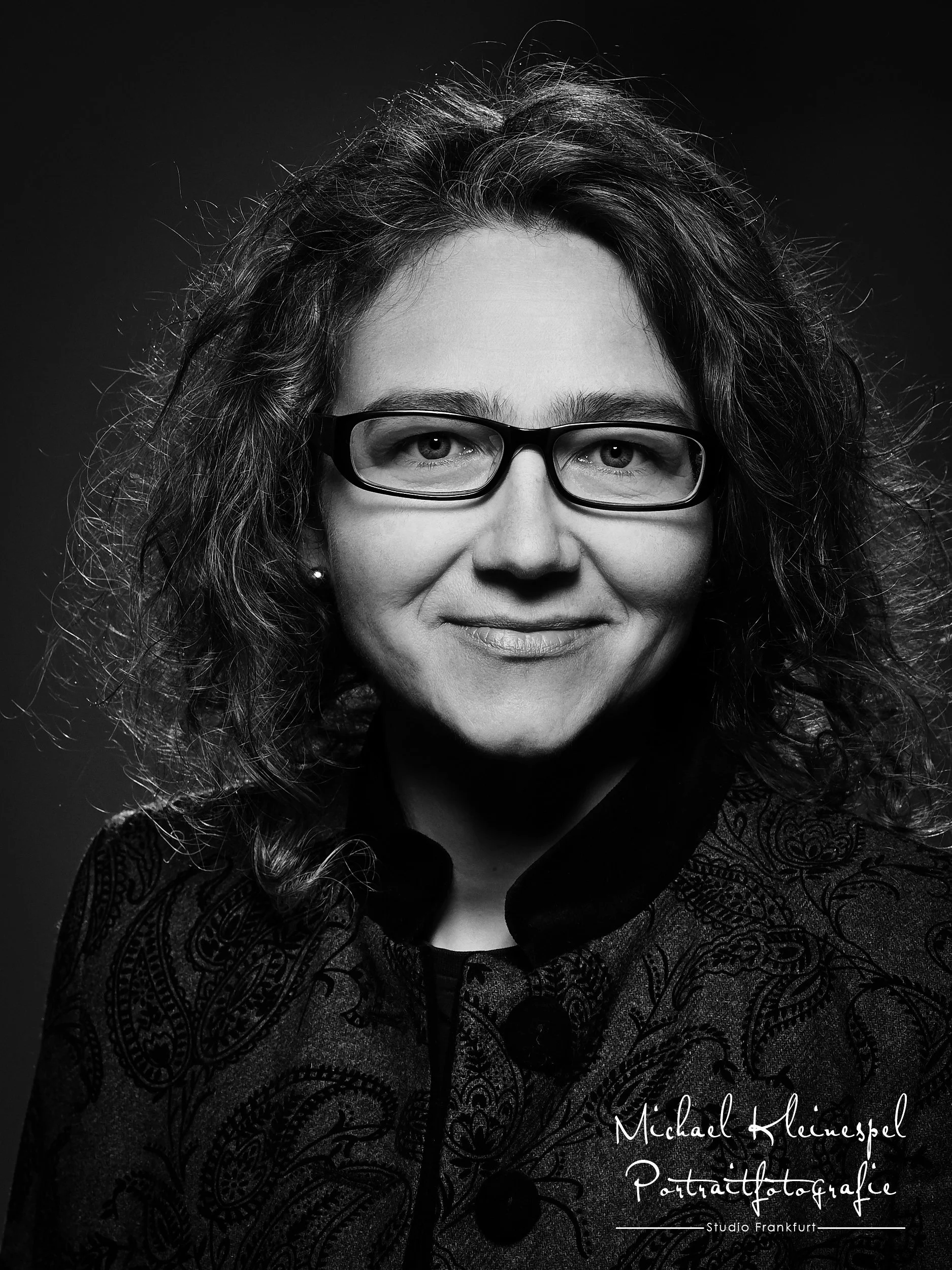 Schwarz-weiß Porträt einer Frau mit lockigem Haar, Brille und einem Lächeln, trägt eine dunkle Jacke mit Paisley-Muster.