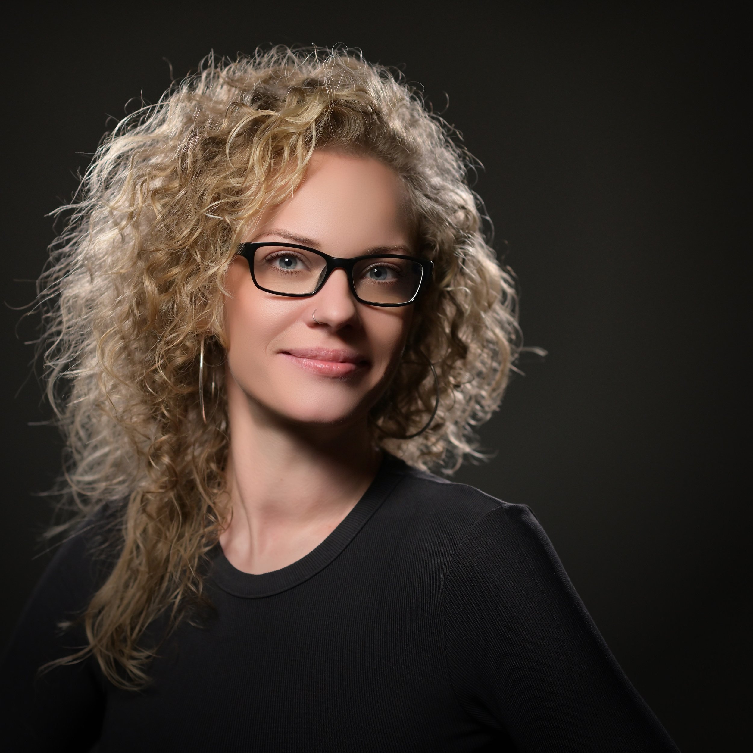 Porträt einer Frau mit lockigem blondem Haar, schwarzen Brillen und dunklem Hemd vor dunklem Hintergrund.