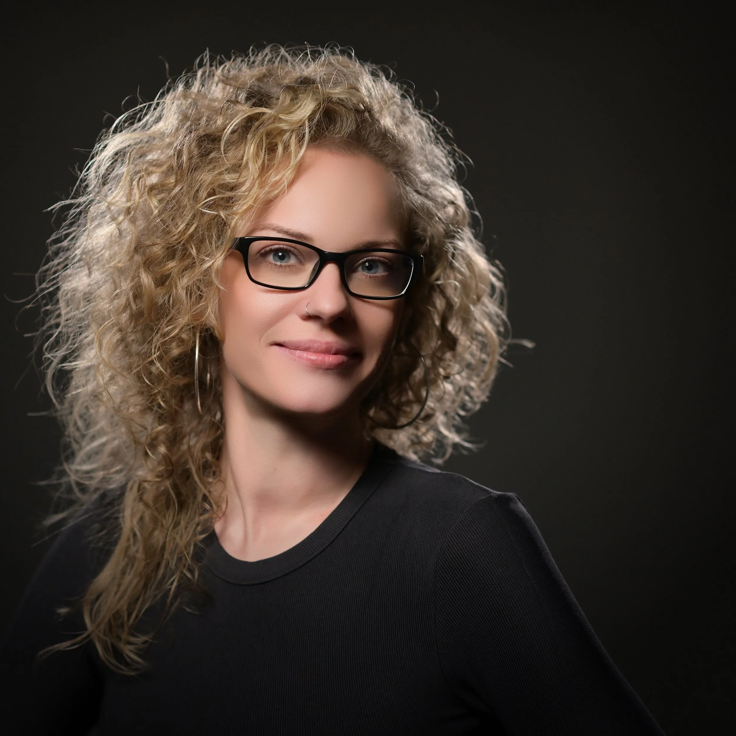 Porträt einer Frau mit lockigem blondem Haar, schwarzen Brillen und dunklem Hemd vor dunklem Hintergrund.