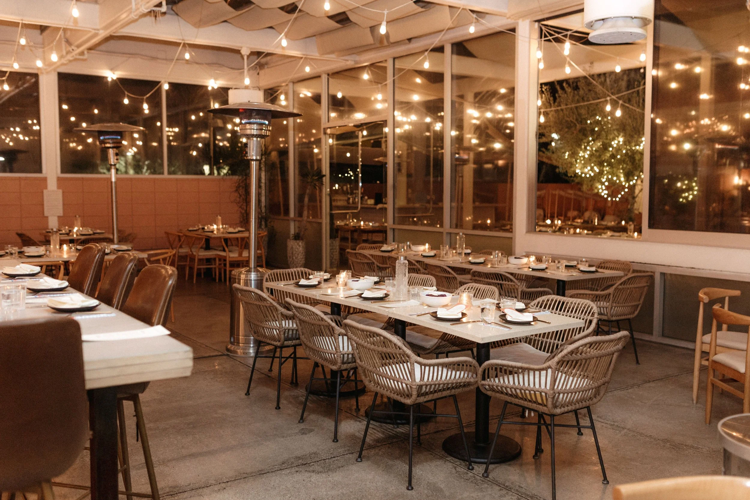 Outdoor restaurant or patio dining area decorated with string lights, with tables set with plates, glasses, and napkins, and surrounded by wicker and wooden chairs. Large windows reflecting string lights create a cozy ambiance.