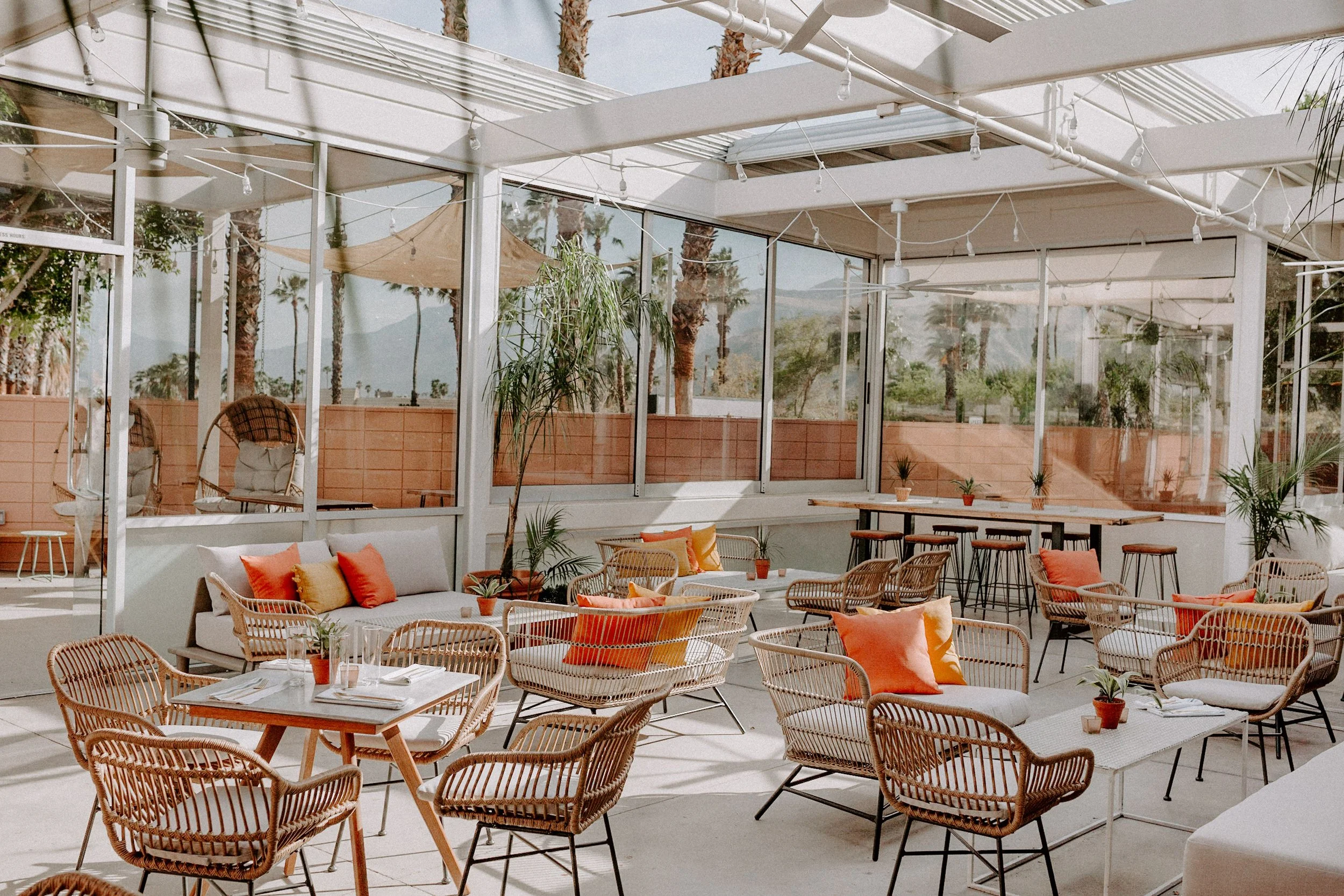 Outdoor patio area with wicker chairs, some with orange and beige cushions, and tables with potted plants. Glass walls reflect palm trees and a mountain view, with string lights hanging from the ceiling.
