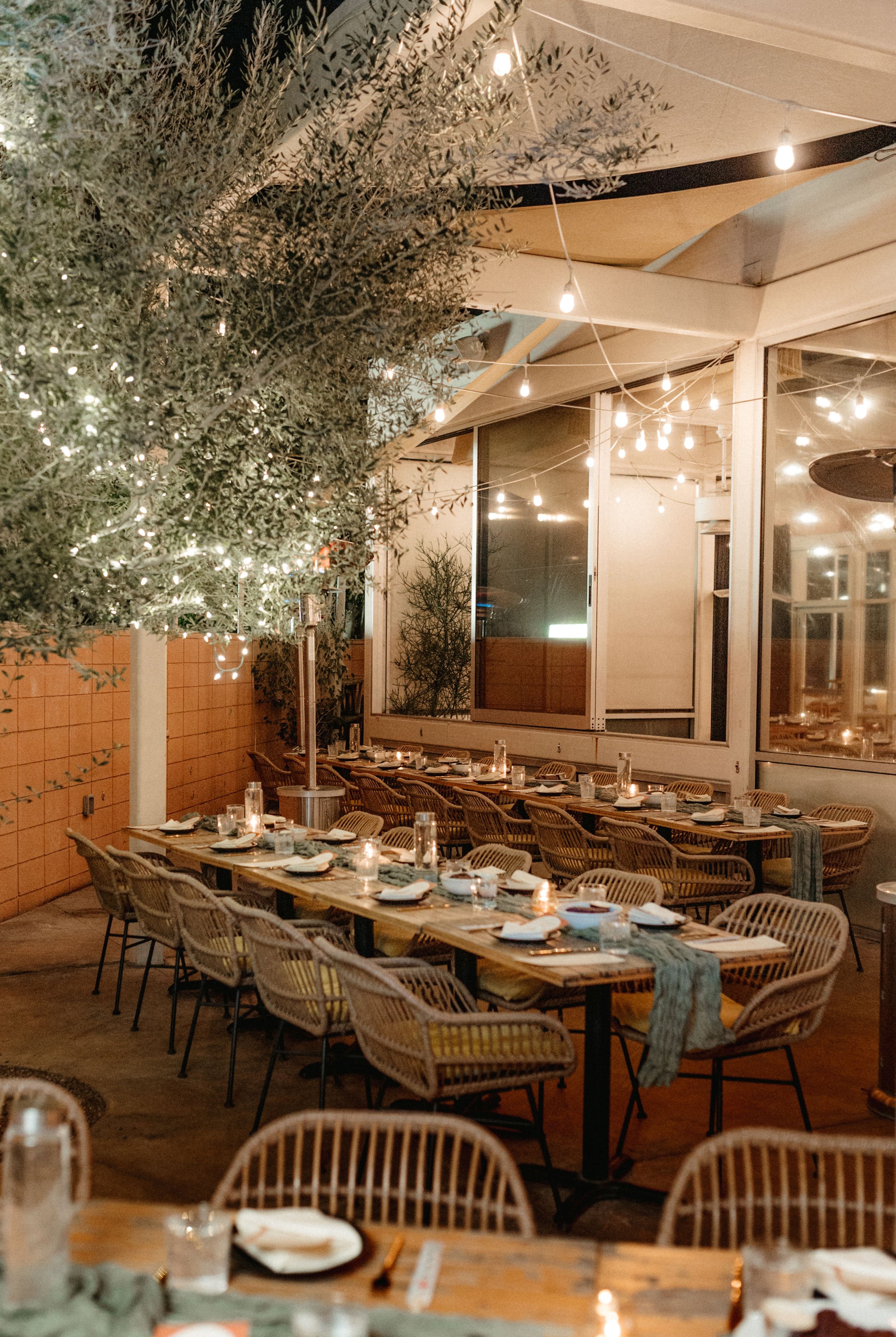 A cozy indoor dining area decorated for a meal with string lights overhead, a long wooden table set with plates, glasses, candles, and napkins, surrounded by wicker chairs.