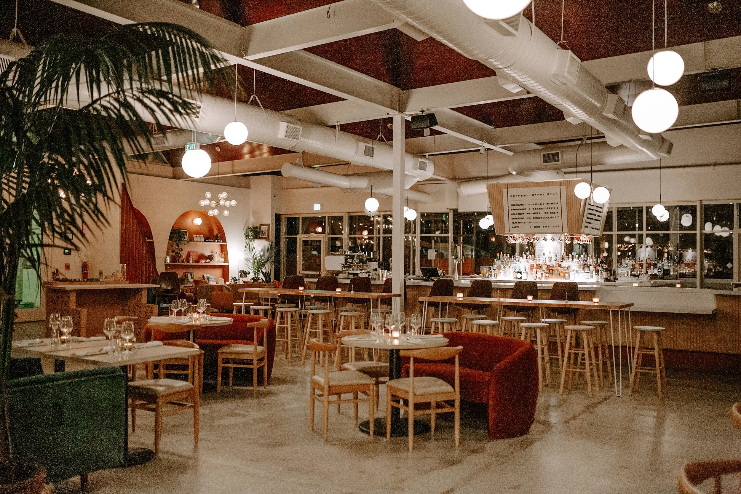 Interior of a restaurant or bar with tables, chairs, and bar counter, illuminated by round hanging lights, with a large menu board visible in the background.