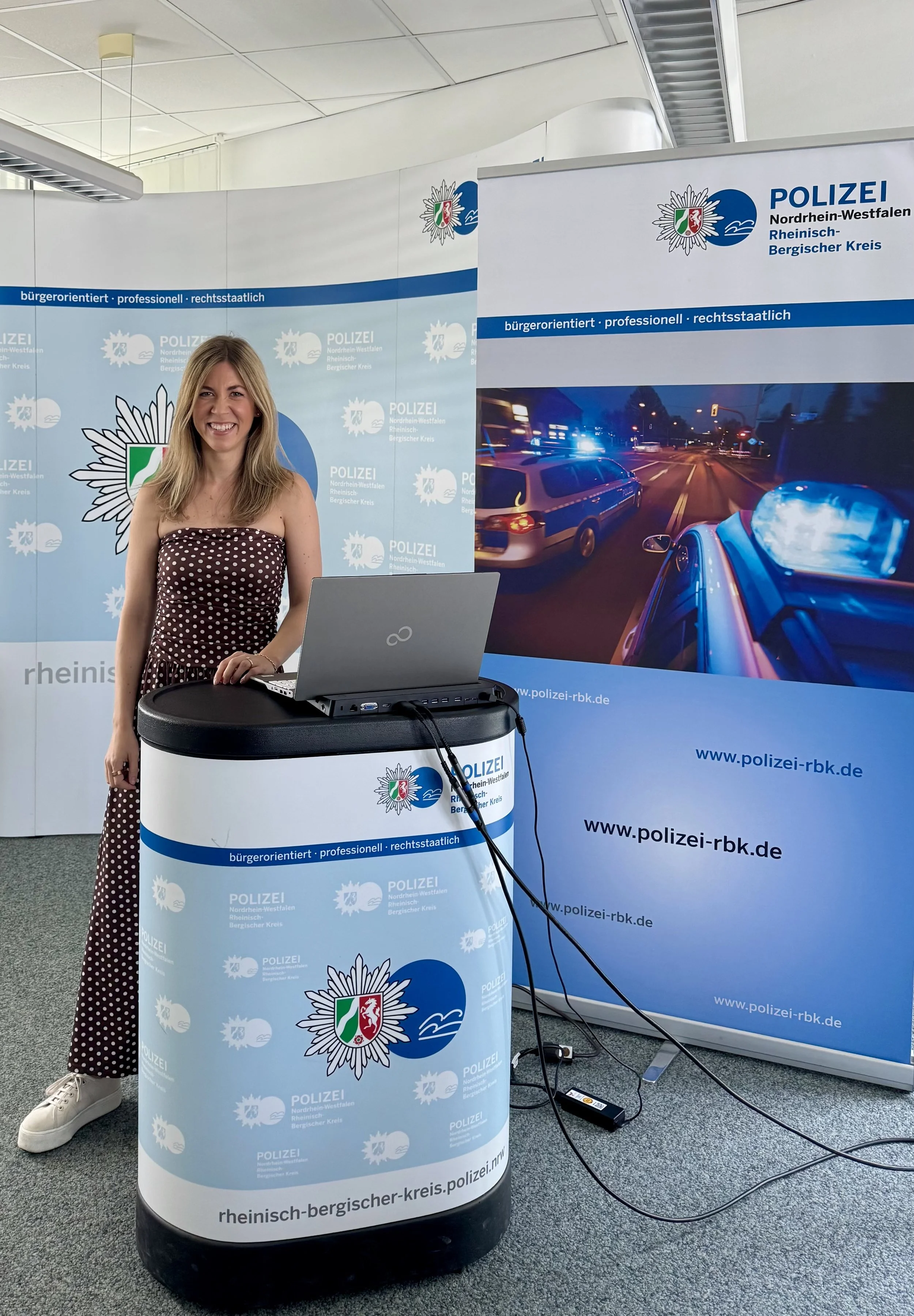 Eine lächelnde junge Frau steht hinter einem runden Info-Stand der Polizei Nordrhein-Westfalen, der mit dem Logo und der Website www.polizei-rbk.de versehen ist. Im Hintergrund befindet sich ein Banner mit einem Polizeifahrzeug bei Nacht und polizeilichen Symbolen und Text.