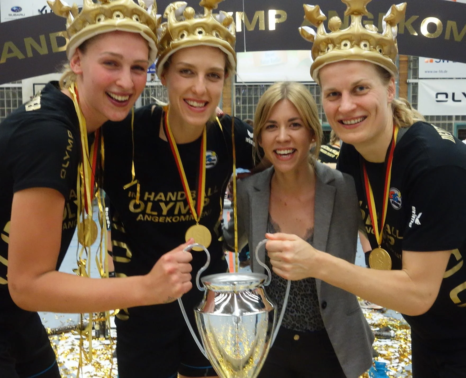 Vier Frauen tragen goldene Kronen und Medaillen und halten eine große silberne Trophäe. Sie lachen und feiern im Hintergrund sind goldene Konfetti und ein Publikum, vermutlich bei einem Sportevent.