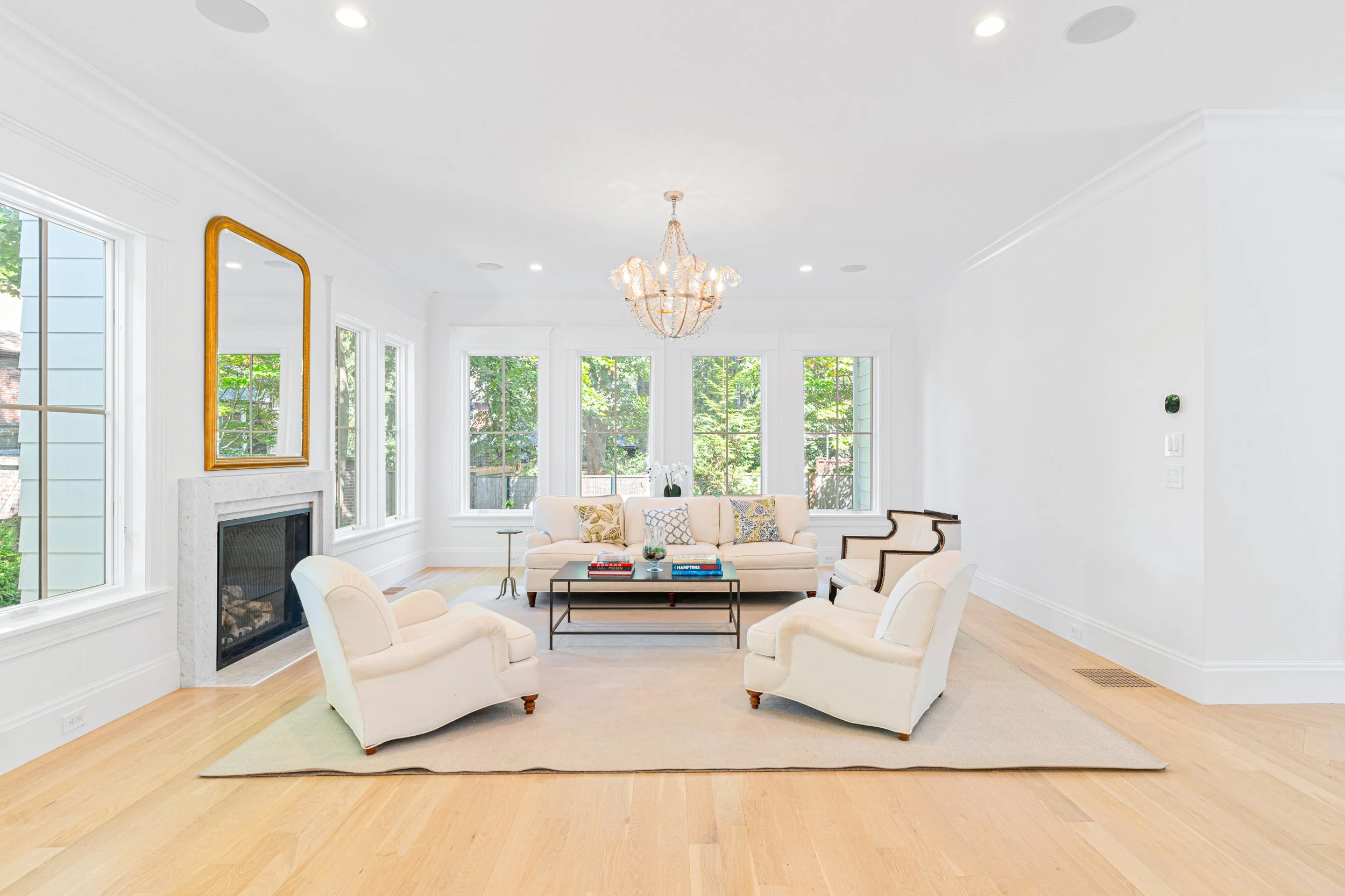 Bright living room in Newton, MA, with white walls, light wood floor, large windows, chandelier, fireplace, and white furniture, including a sofa and armchairs.