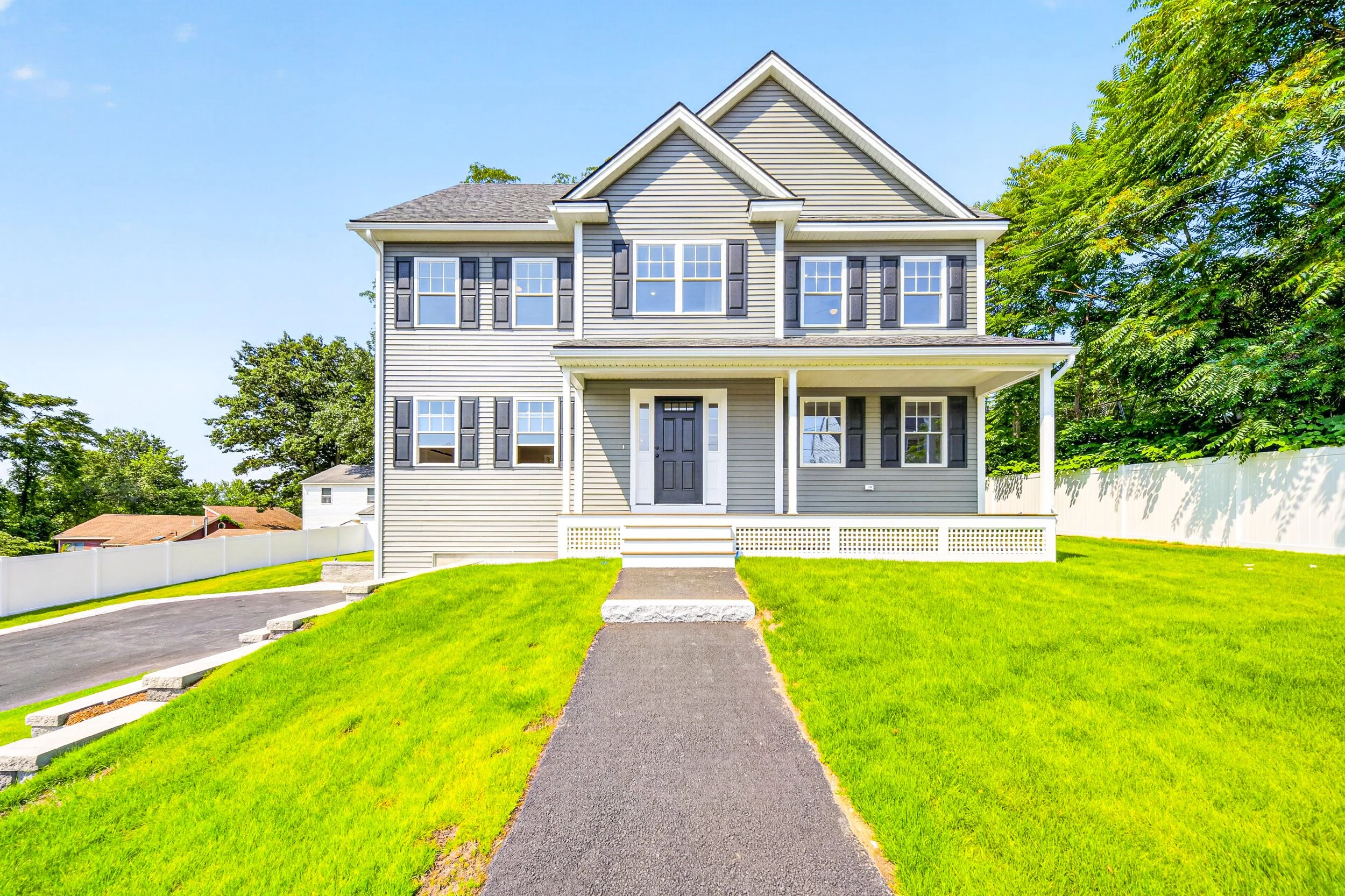 A two-story gray house in Billerica, MA, with black shutters, white trim, and a covered front porch. It has a black front door and a small sidewalk leading to the steps, surrounded by a well-manicured lawn, trees, and a white fence in the background.