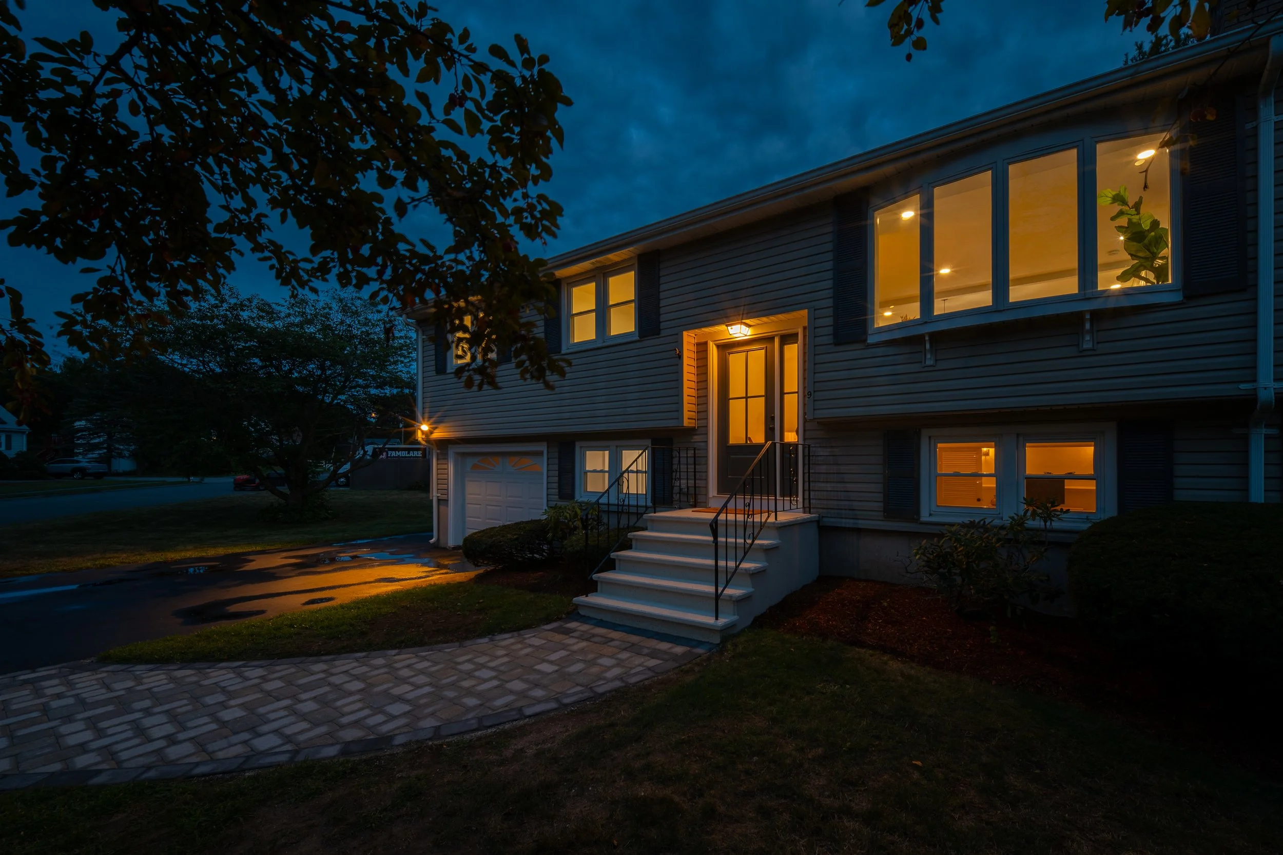 A two-story house in Billerica, MA, with illuminated windows and front porch light during dusk. The house has beige siding, a set of stairs leading to the front door, and a white garage door.