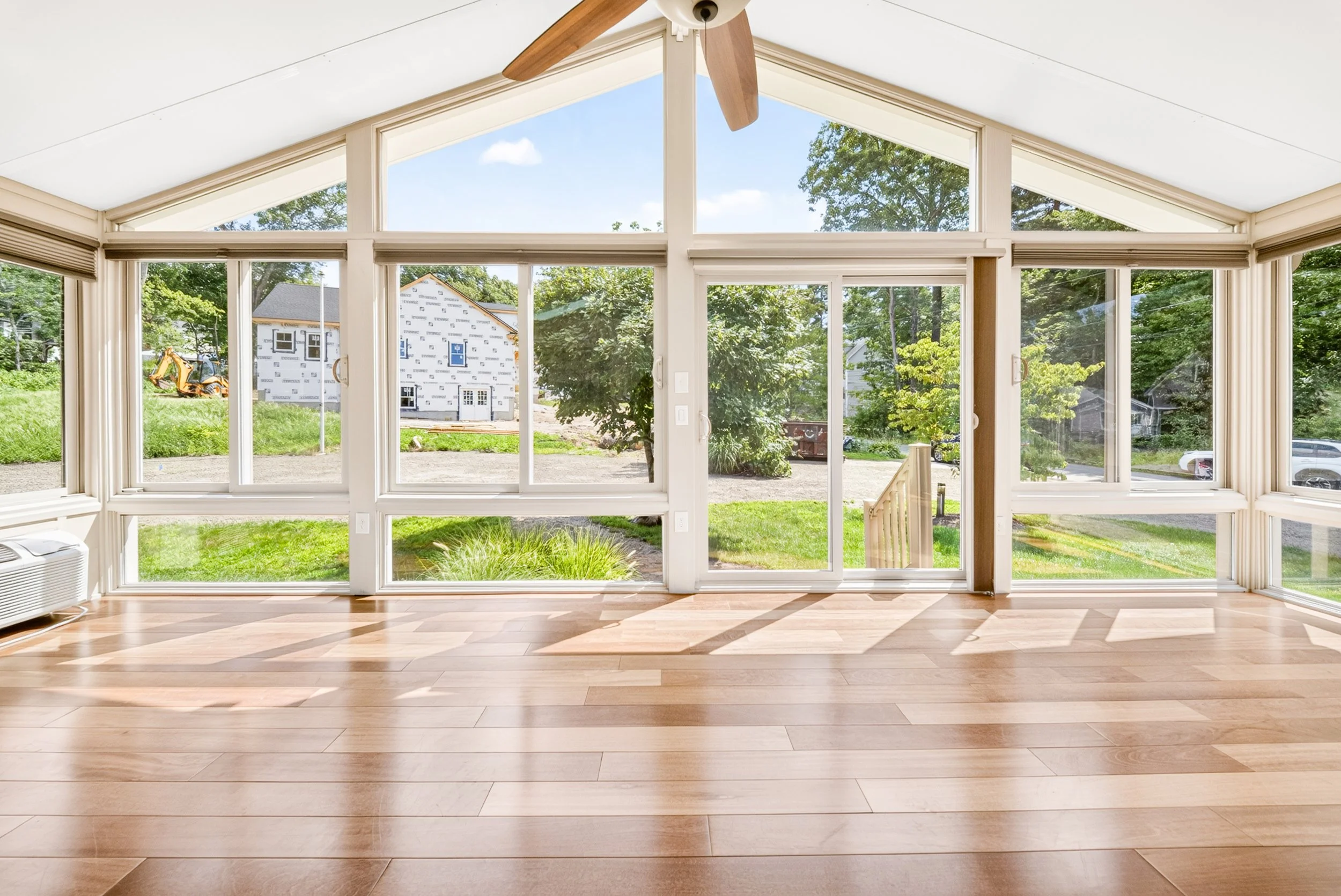 View from a house in Billerica, MA, inside a sunroom with large windows and a sliding glass door. Outside, there are green trees, a grassy yard, a construction site with a partially built house, and a small excavator. The sky is clear with clouds.
