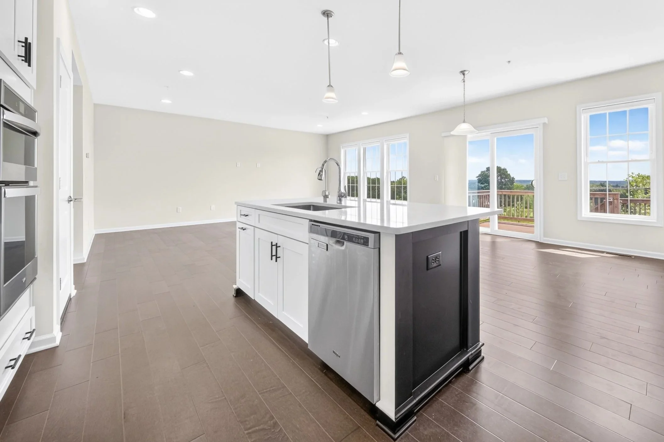 Modern open-plan kitchen in Hopkinton, MA, with white island, stainless steel sink, hardwood floors, large windows, and a view of trees and sky outside.