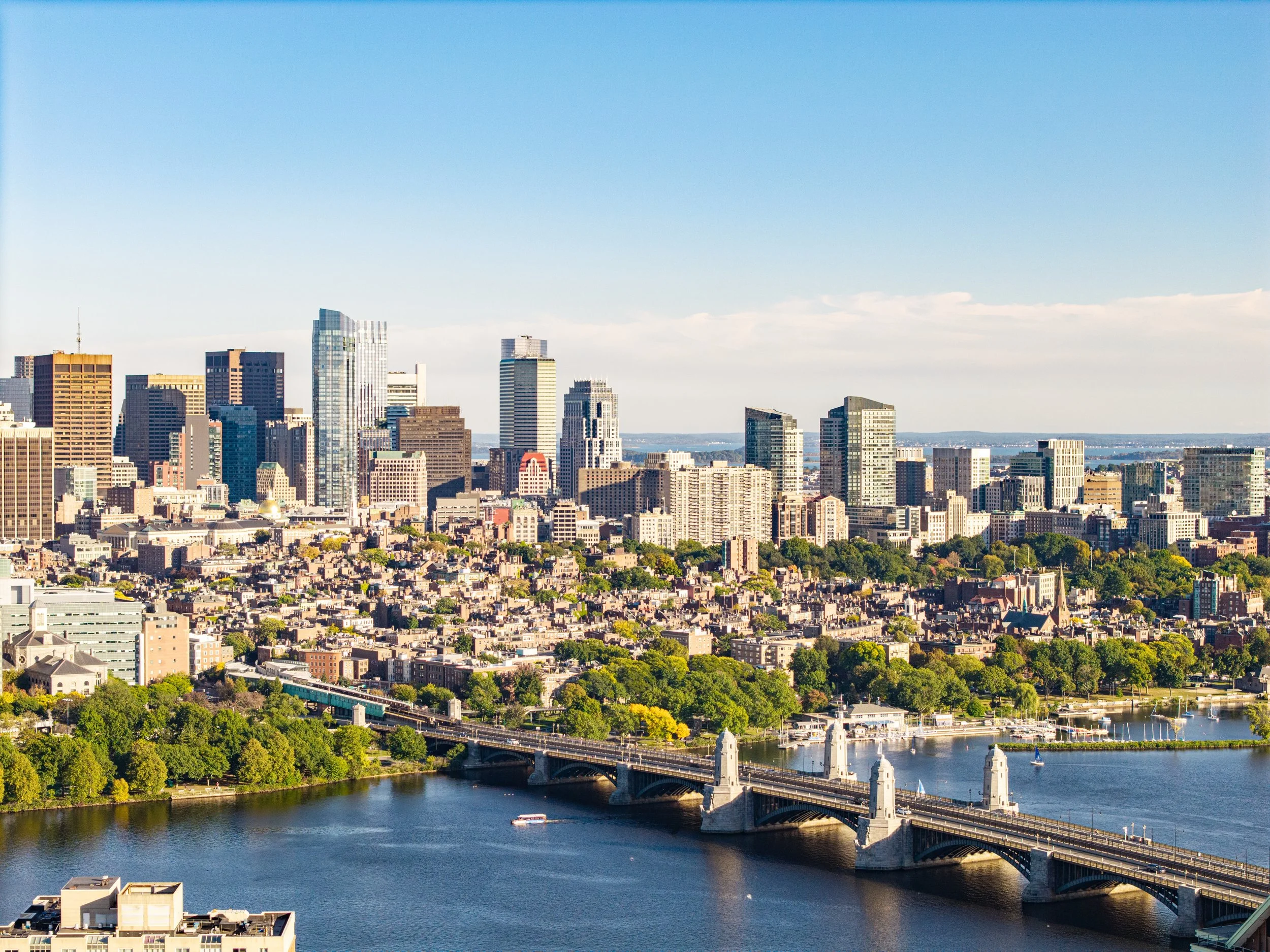 A Boston, MA skyline with tall modern buildings, a river with a bridge, and lush green trees.