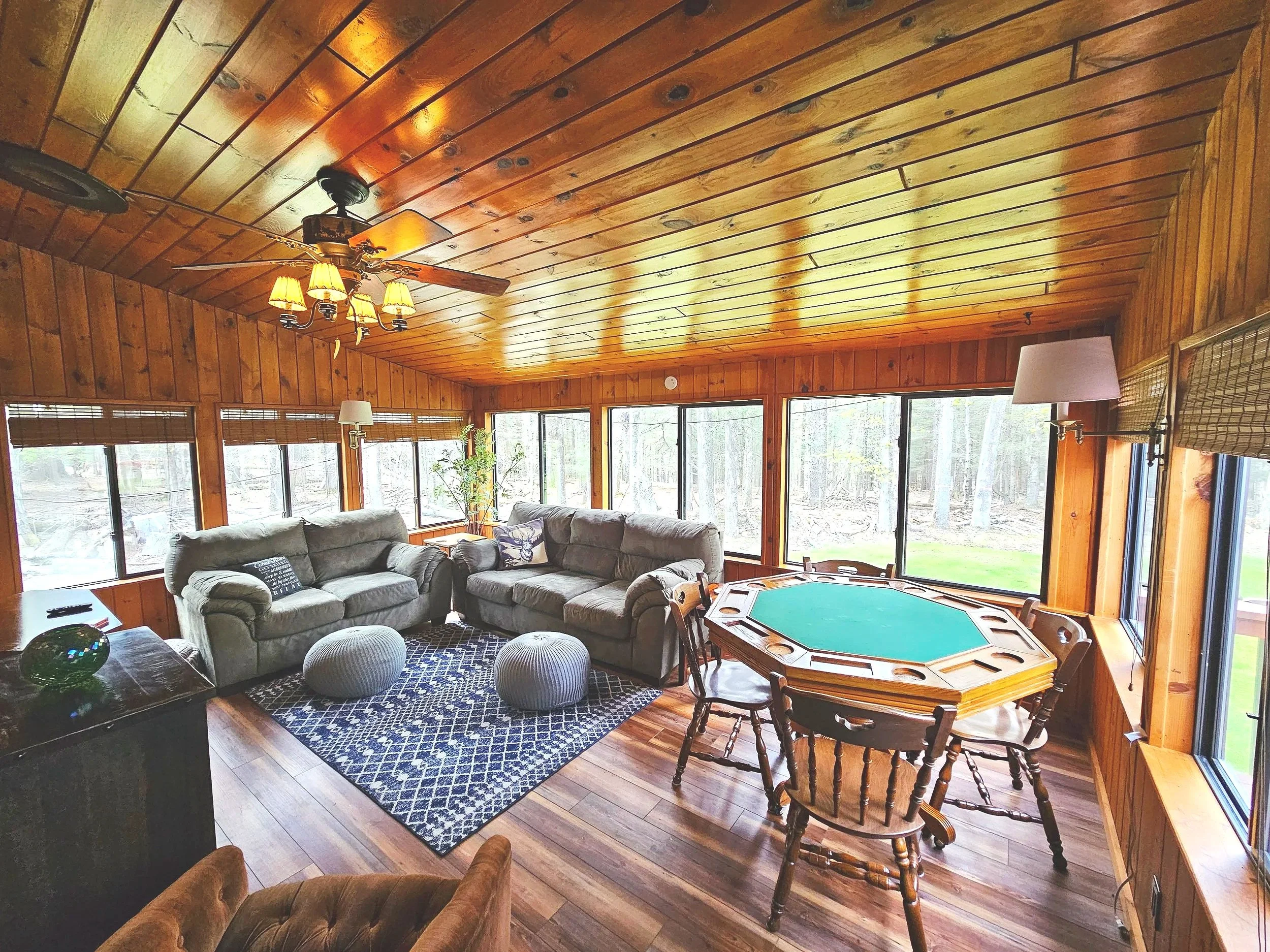 A cozy living room with wood-paneled walls and ceiling, large windows bringing in natural light, 2 comfy sofas, and blackjack table.