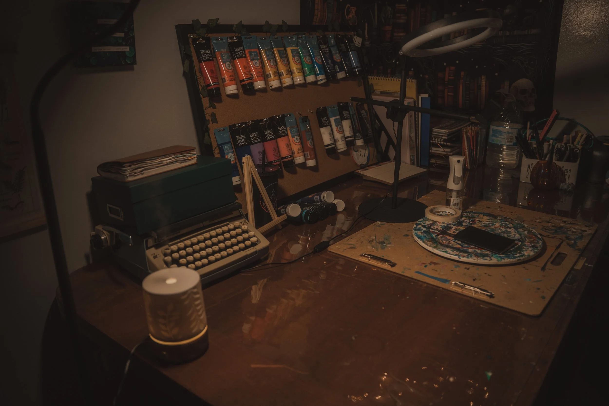 A typewriter, a corkboard of hanging acrylic paint, some other art supplies such as tape, pens, etc. are all arranged on a big brown table.