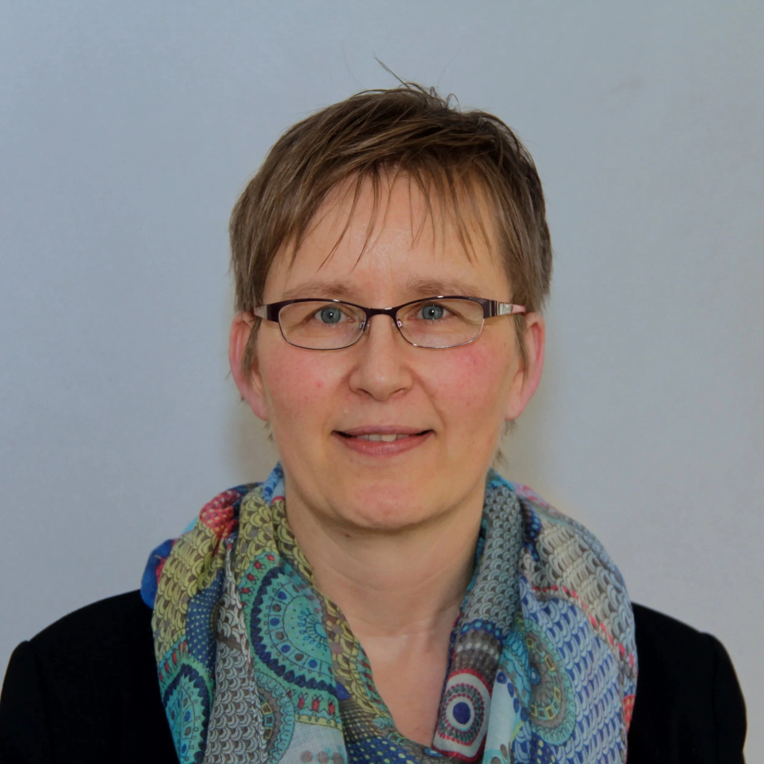 Porträt einer Frau mit Brille, kurzen braunen Haaren, schwarzem Oberteil und buntem Schal vor grauem Hintergrund.