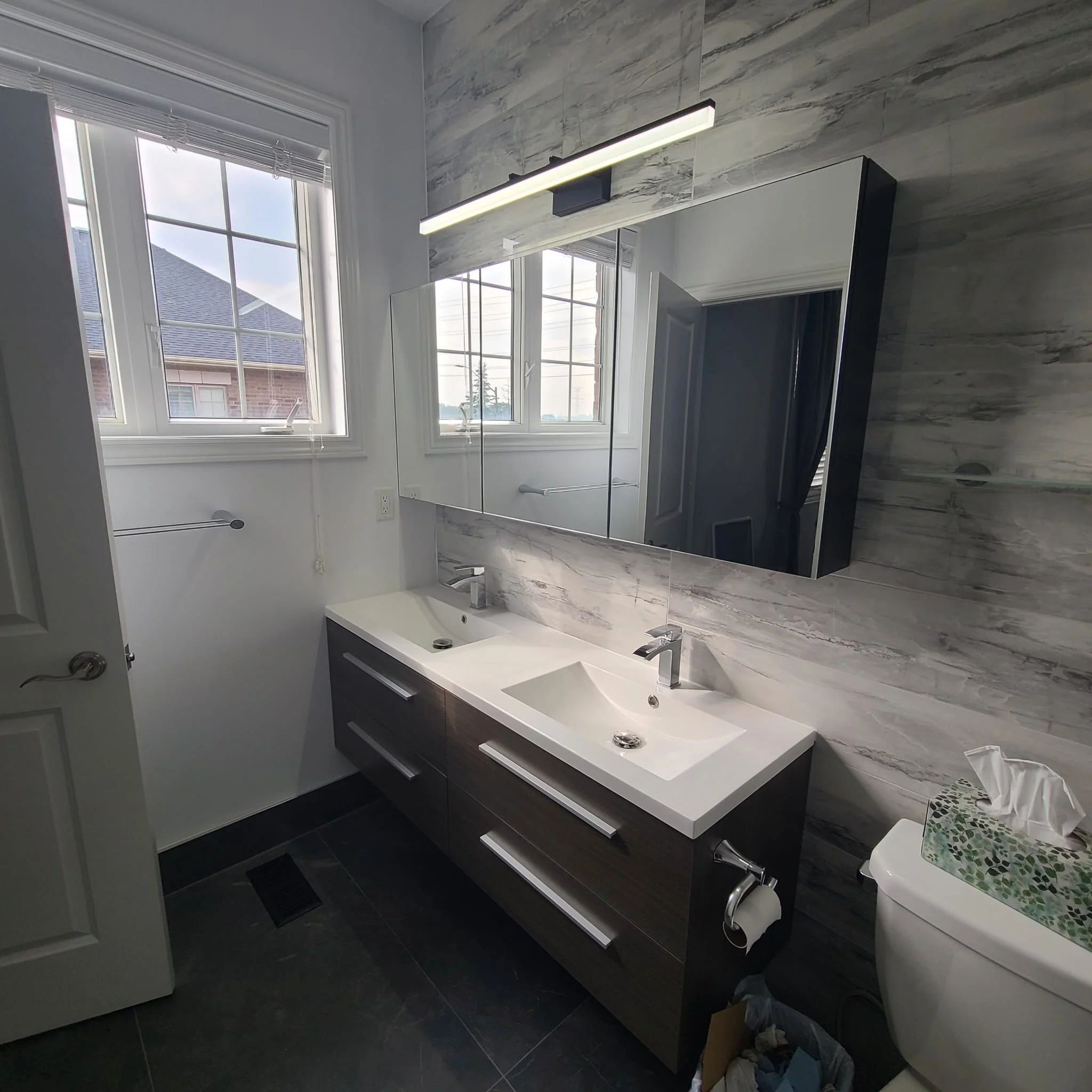 Modern bathroom with dual sinks, large mirror, gray marble wall, window with blinds, white walls, dark cabinet, and a toilet with a box of tissues on top.