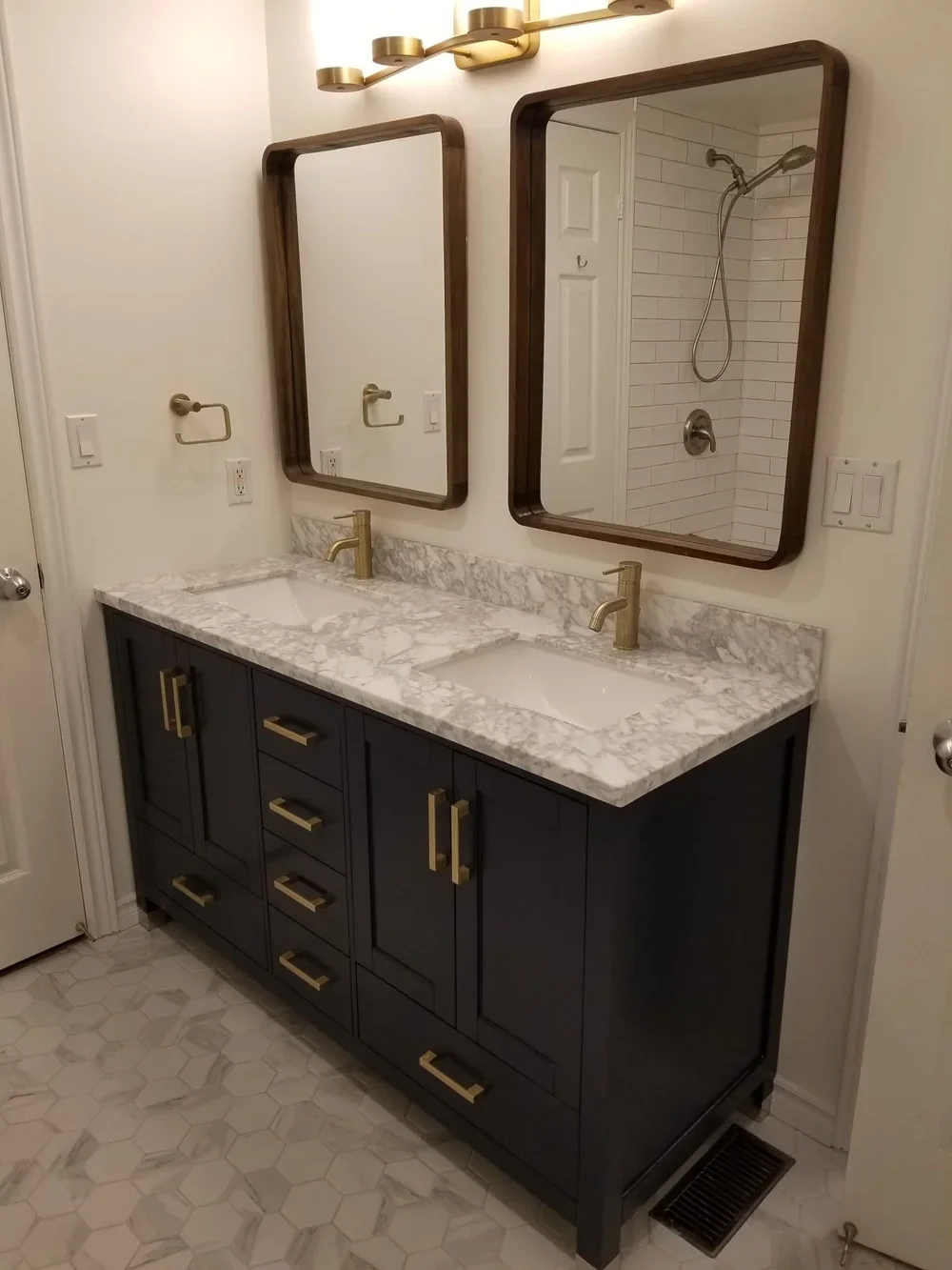 Double bathroom vanity with marble top, two mirrors, gold fixtures, and navy blue cabinets, with a shower visible in the mirror reflection.