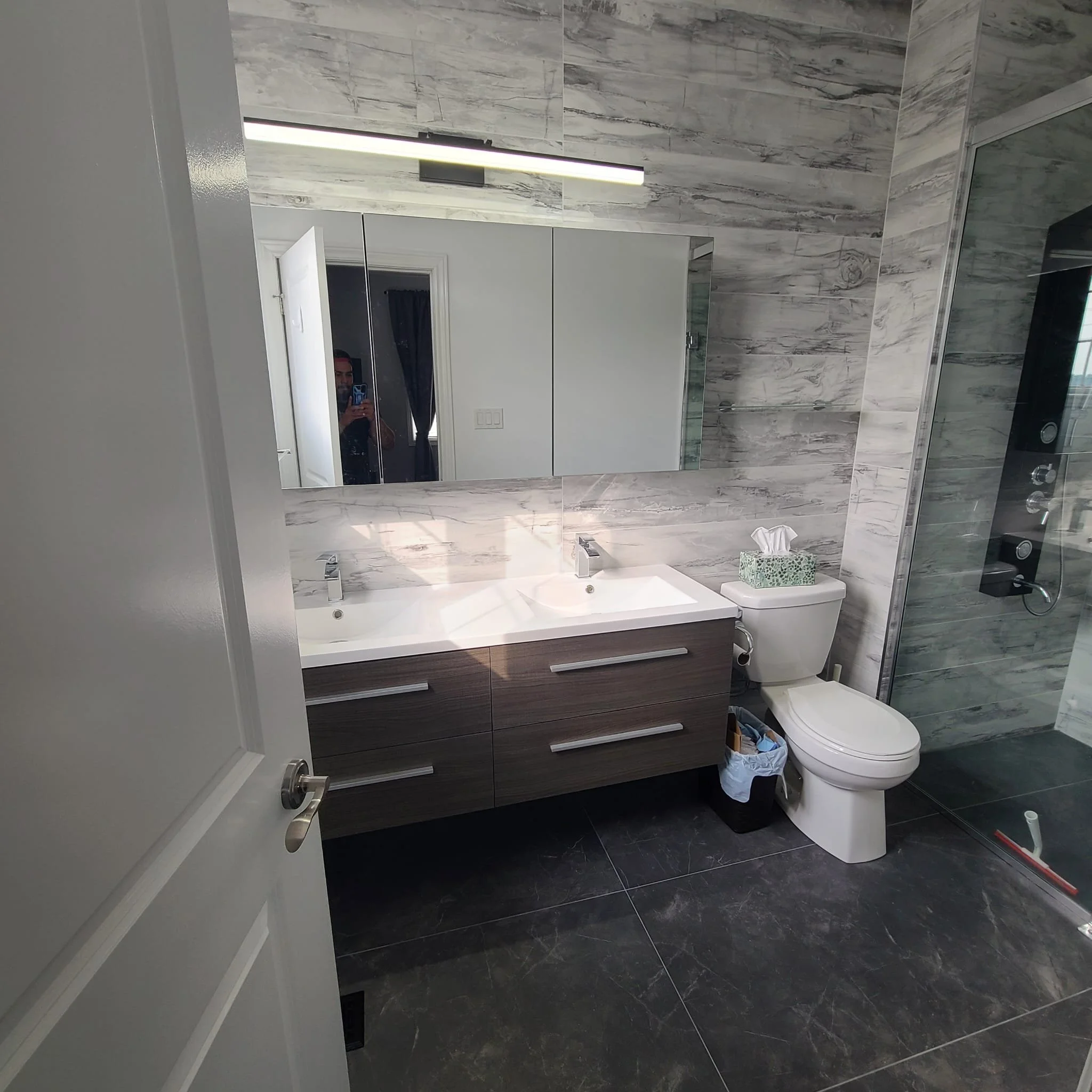Modern bathroom with marble walls, a large mirror, double vanity sinks with dark wood cabinets, a toilet with a tissue box on top, and a glass shower enclosure with a black panel of controls.