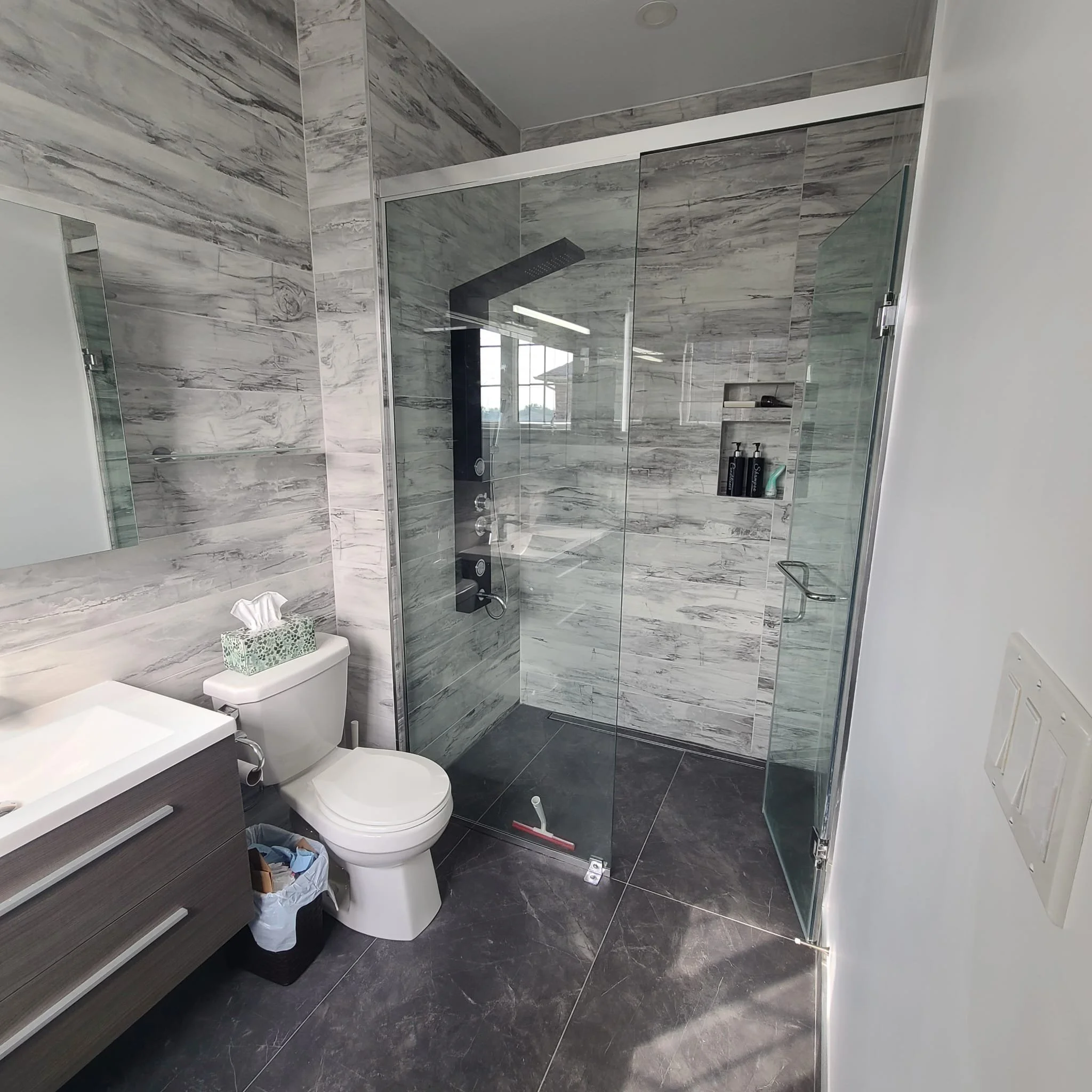 A modern bathroom with a glass-enclosed shower featuring a black vertical shower panel, a white toilet with a tissue box on top, a dark wood vanity with a white sink, and large gray marble tiles on the walls and floor.