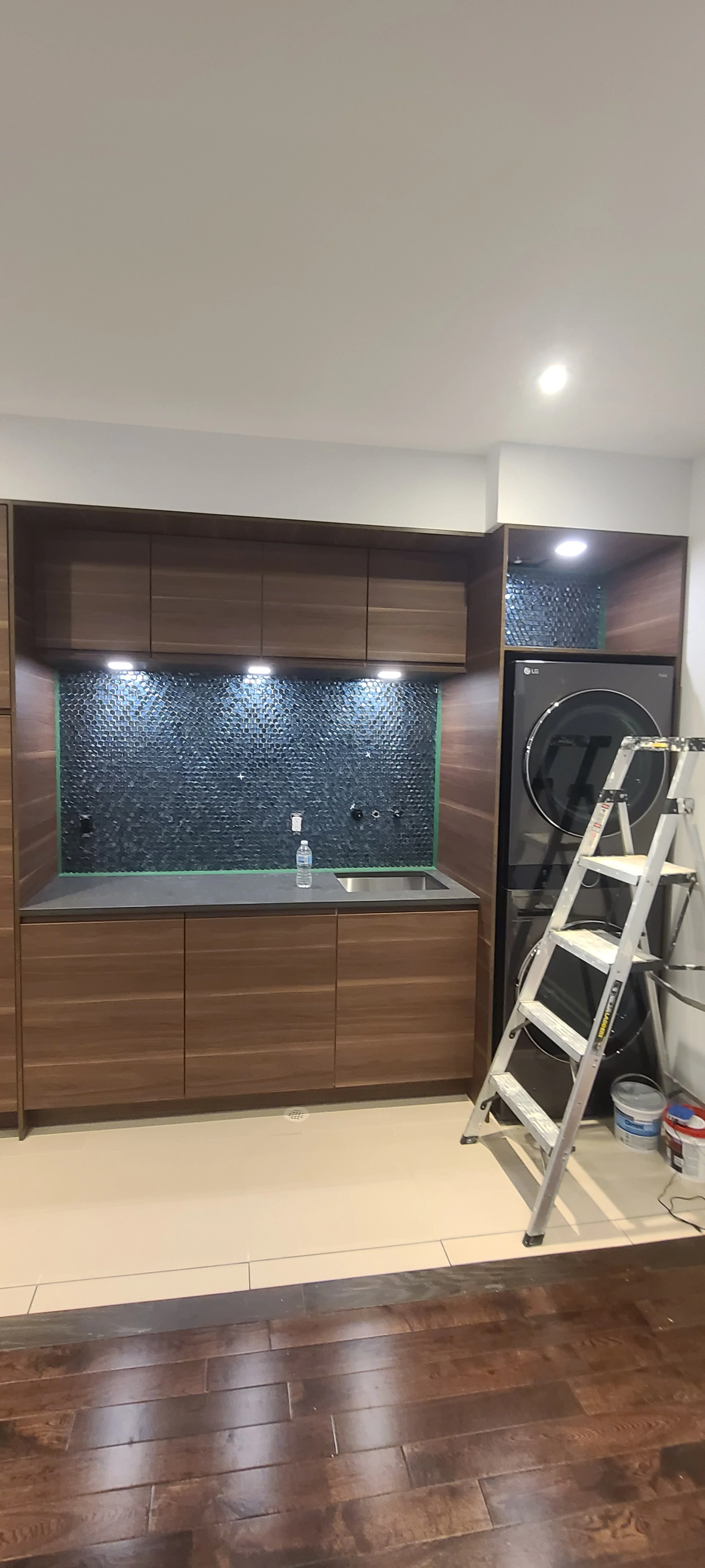 Kitchen with dark wood cabinets, black tiled backsplash, a small sink, a water bottle, a step ladder, and a washer-dryer unit in the corner.