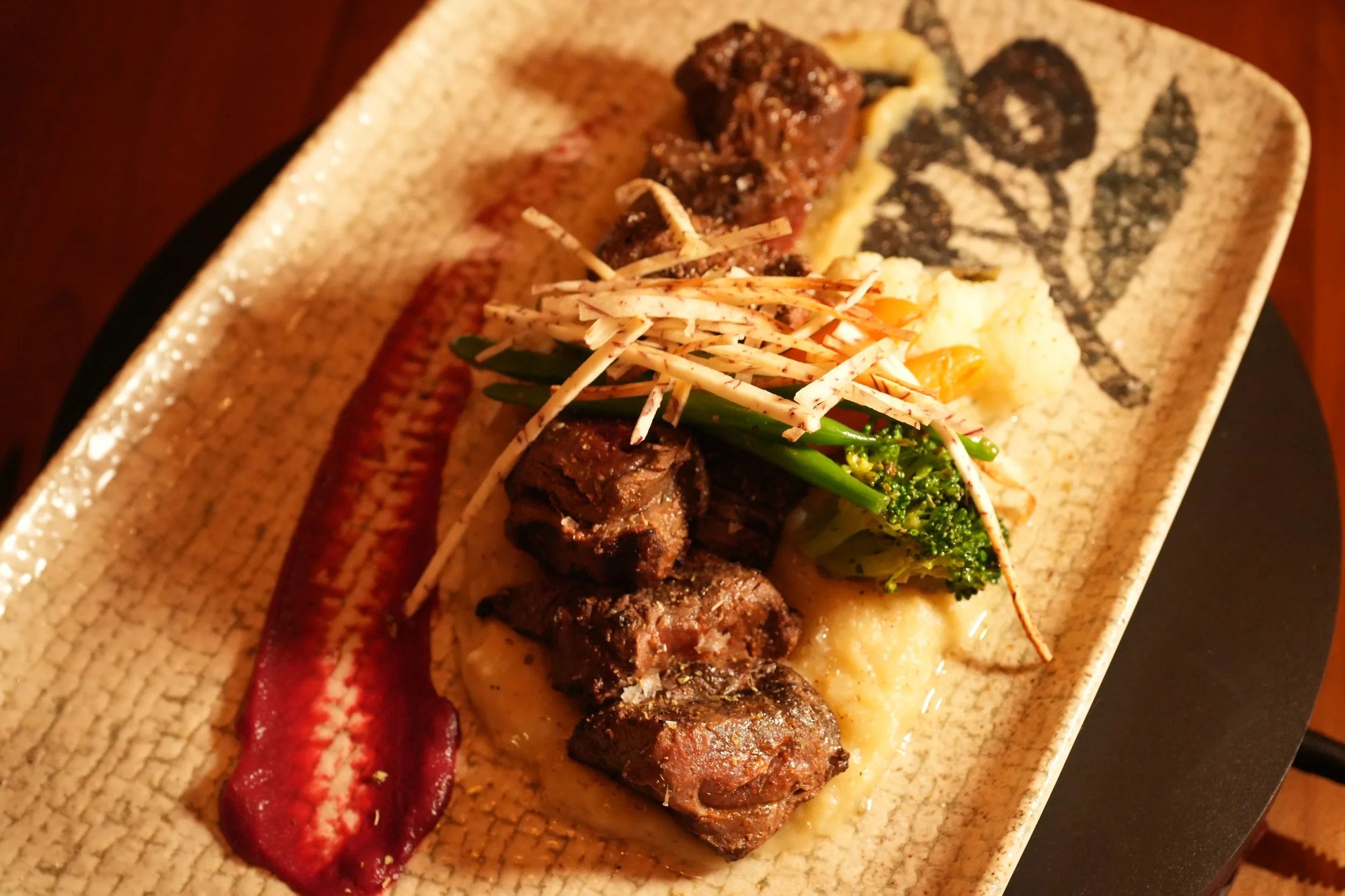 A rectangular ceramic plate with grilled meat, mashed potatoes, broccoli, and shredded vegetables on top, with a red sauce painted on the plate.