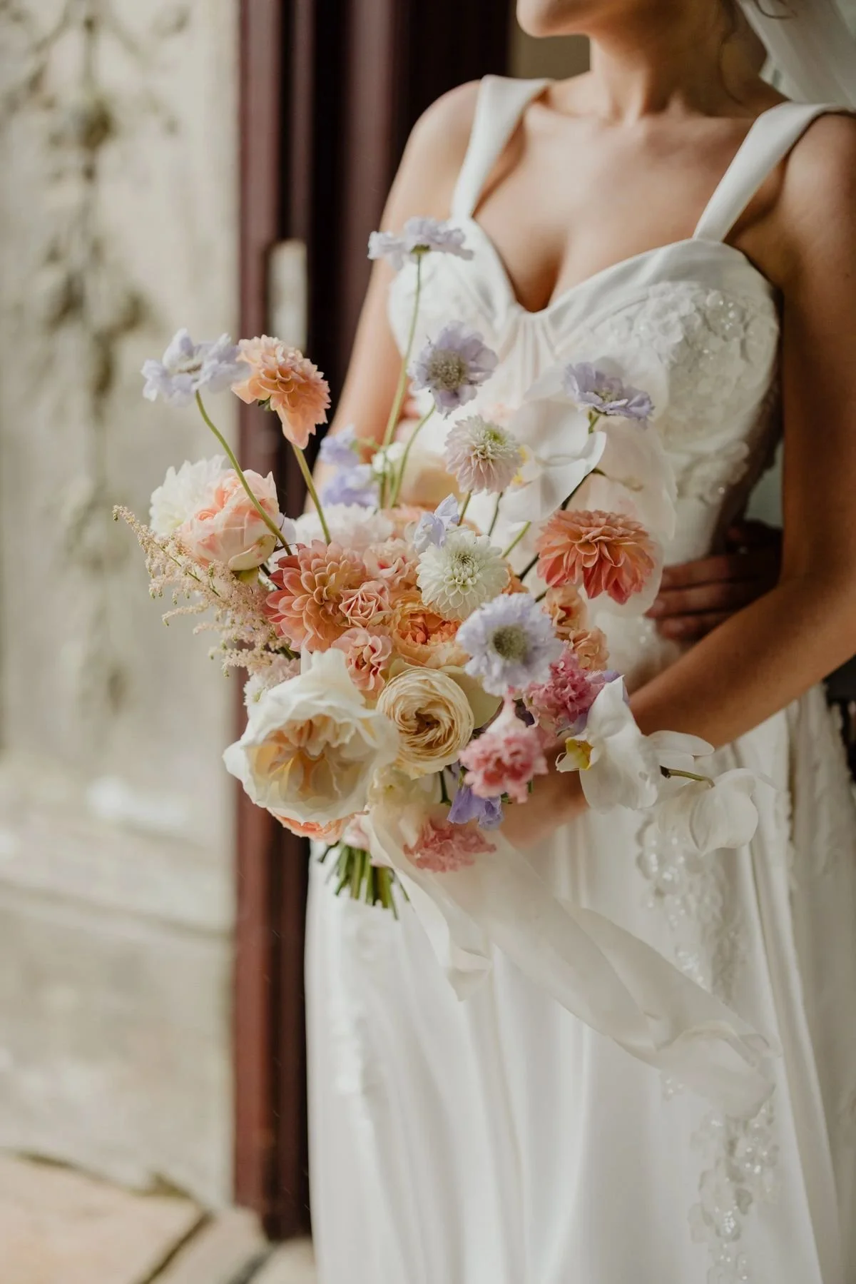 A bride in a white wedding dress holding a bouquet of pastel-colored flowers, standing indoors with a blurred background.