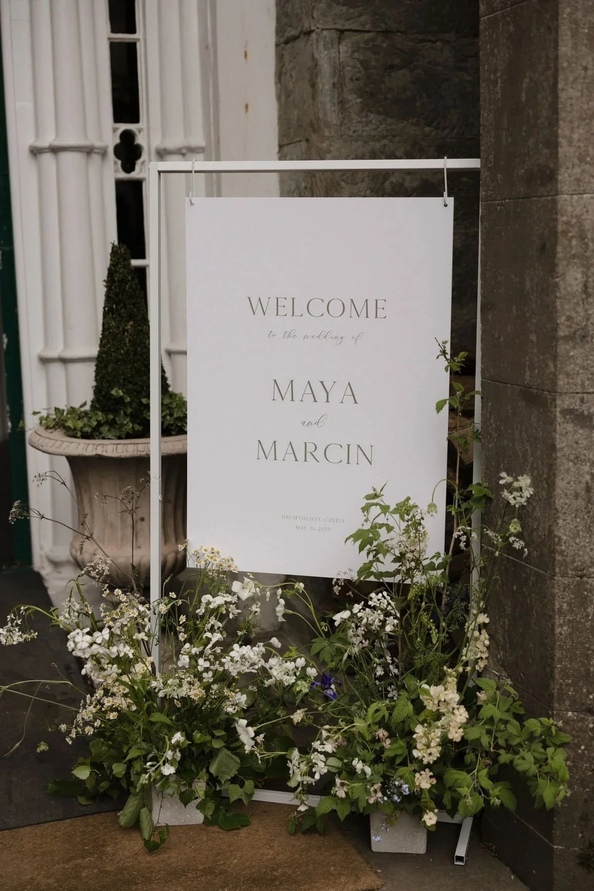 A wedding welcome sign on a metal stand, surrounded by floral arrangements with white flowers and greenery, located outside a building with stone and white wooden architectural details.
