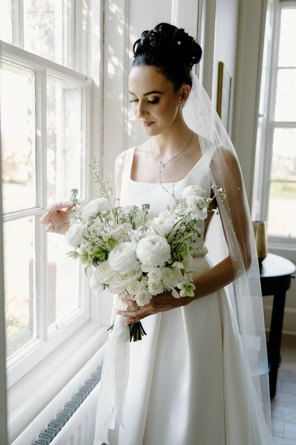 Bride in wedding dress holding a bouquet of white flowers near a window.