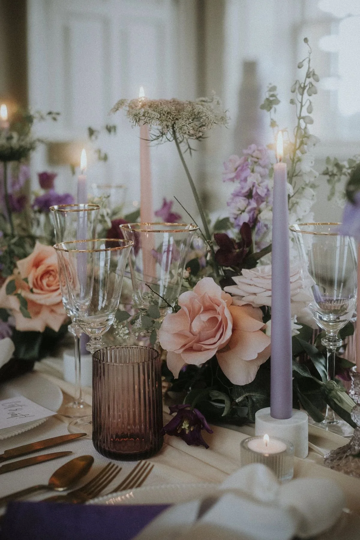 Elegant table setting with pink and purple flowers, gold-rimmed glassware, and lit candles.