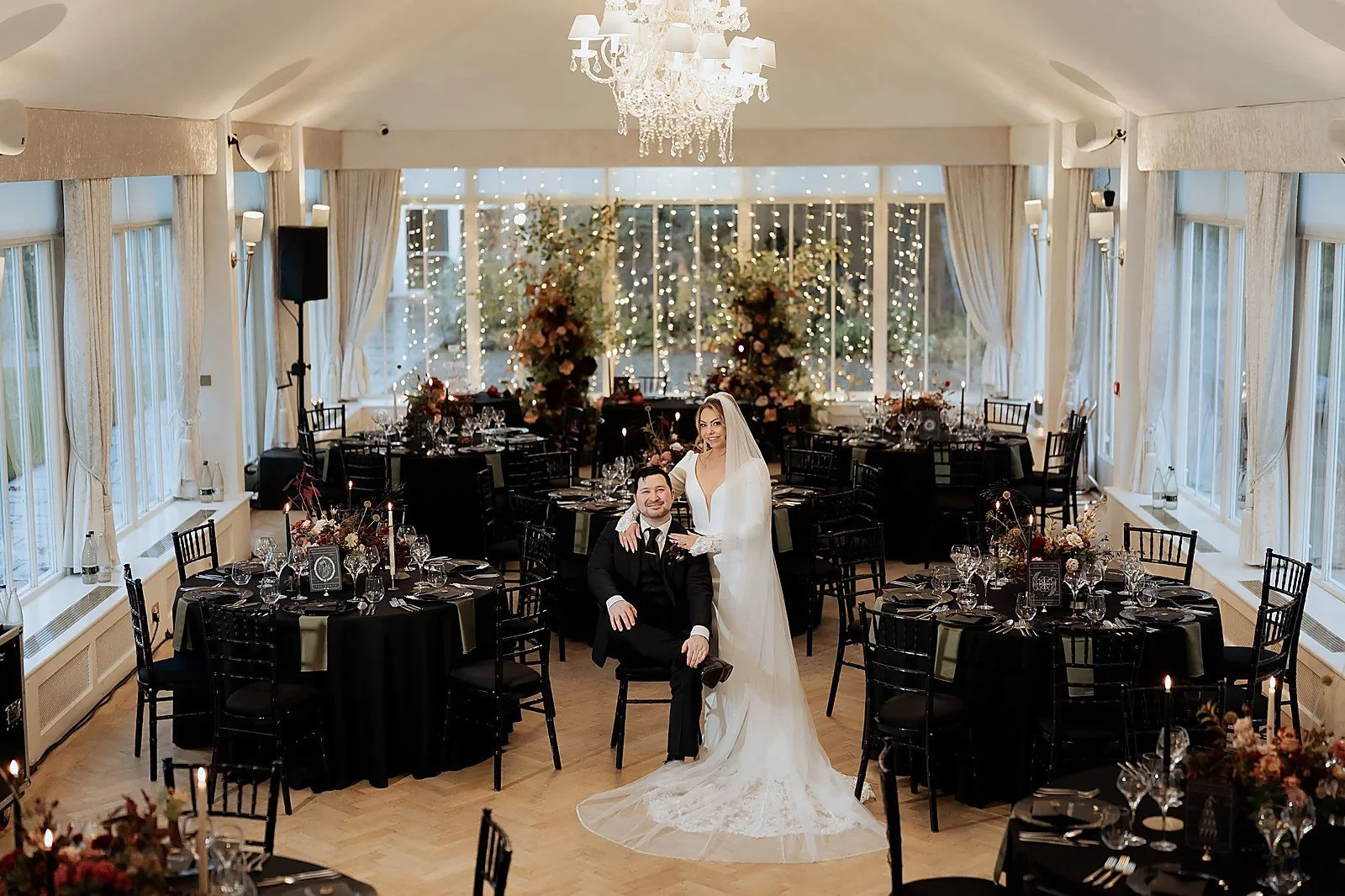 A bride and groom at their wedding reception in a decorated banquet hall with floral arrangements and string lights.